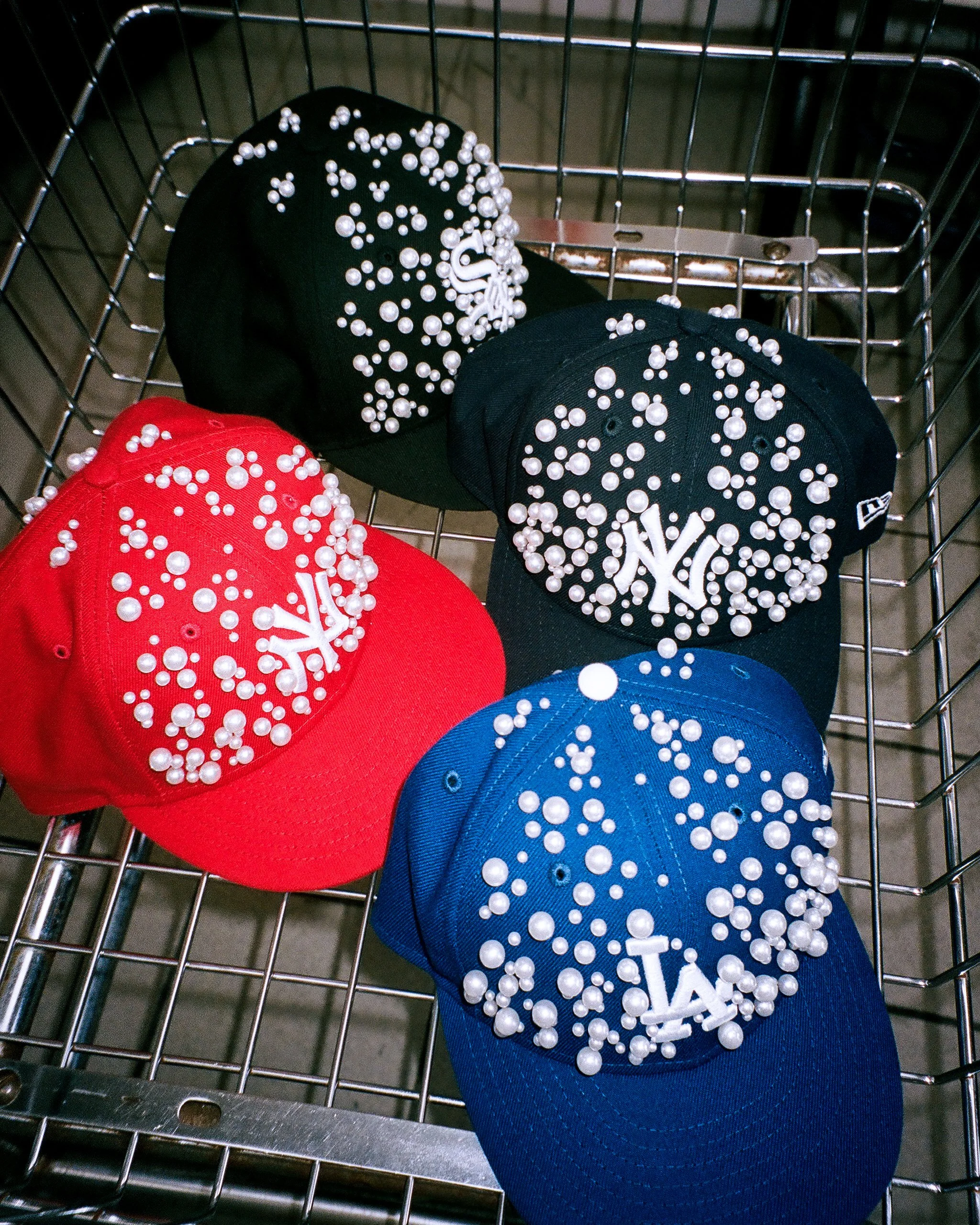 Four New York Yankees baseball caps decorated with white pearls, in red, blue, black, and dark green, placed in a shopping cart.