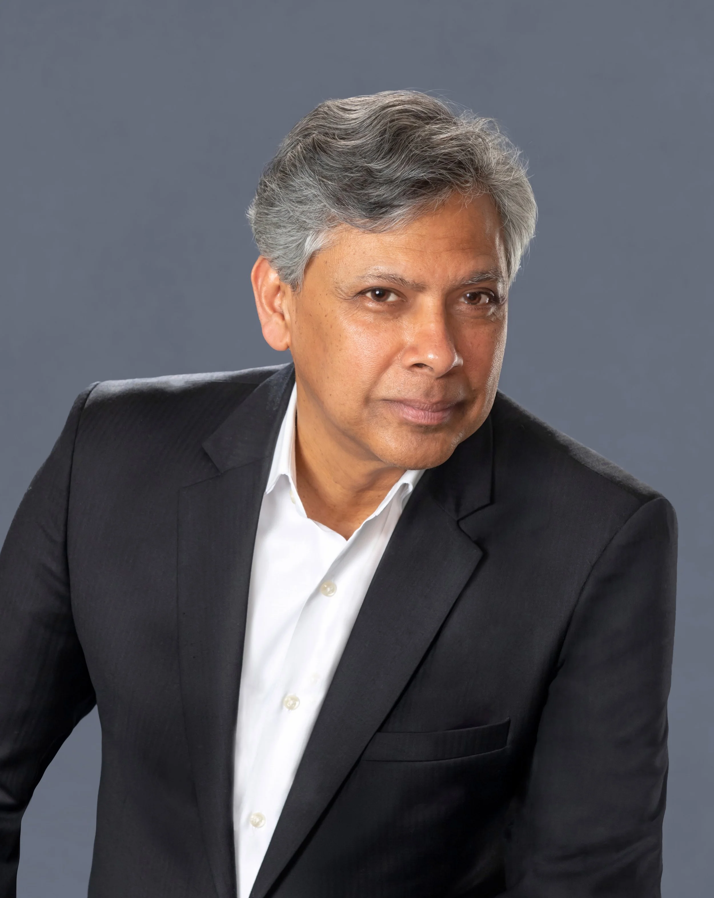 Portrait of a middle-aged man with gray hair wearing a black suit and white shirt, looking confidently at the camera against a gray background.