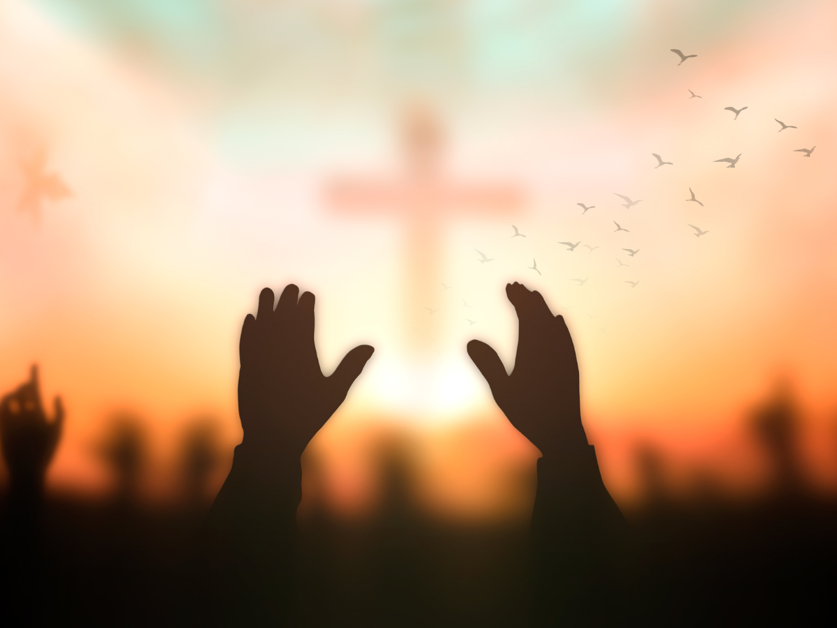 Silhouettes of hands raised in worship against a sunset with a cross and flying birds in the background.