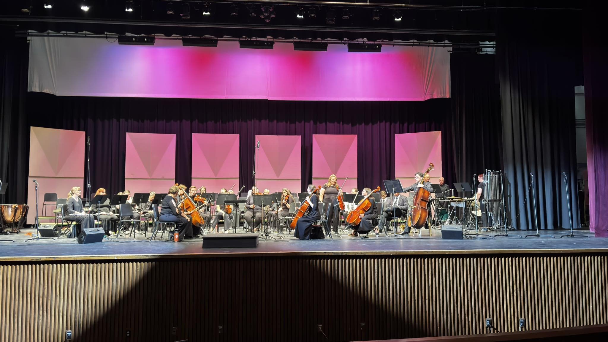 Orchestra on stage with musicians playing instruments under pink and purple lighting with black curtains in the background.
