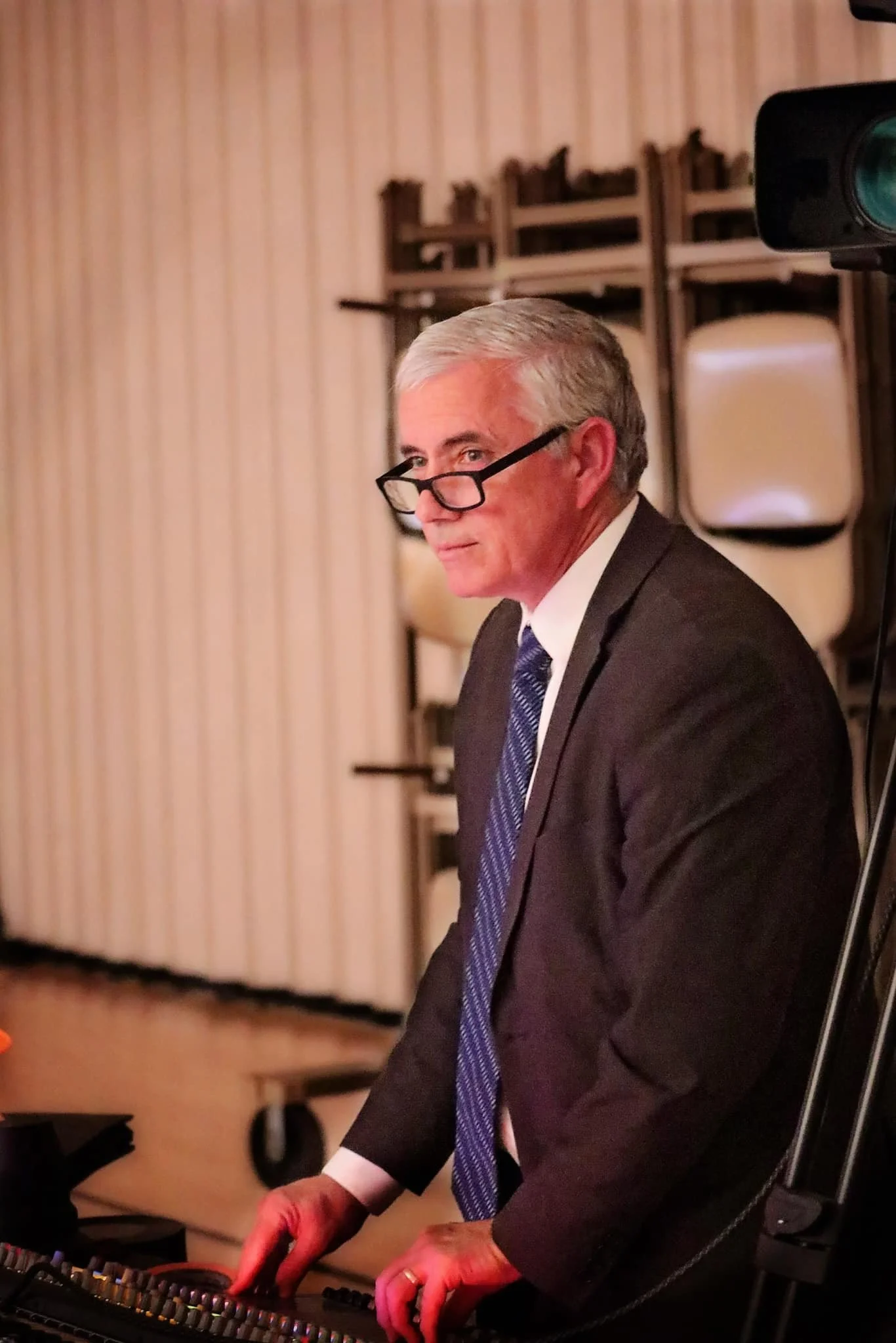 A middle-aged man with gray hair and black glasses, wearing a dark suit with a white shirt and blue tie, stands at a DJ console or electronic music equipment, looking to the side. The background features stacked chairs and a curtain.