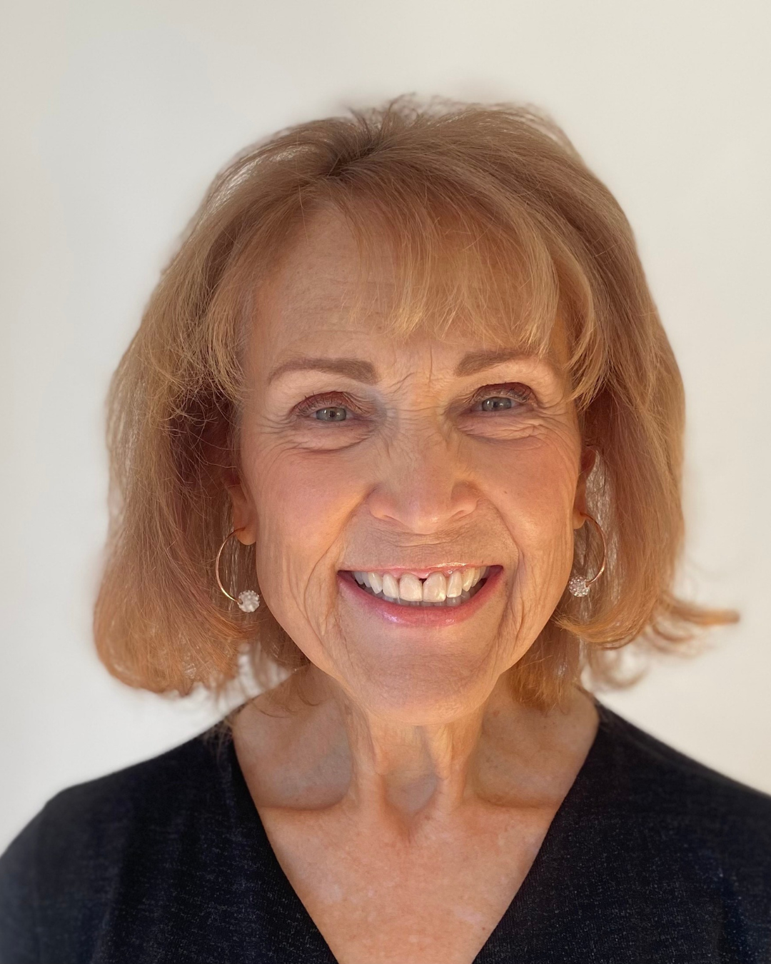 A smiling older woman with short, light brown hair, wearing earrings and a black top.