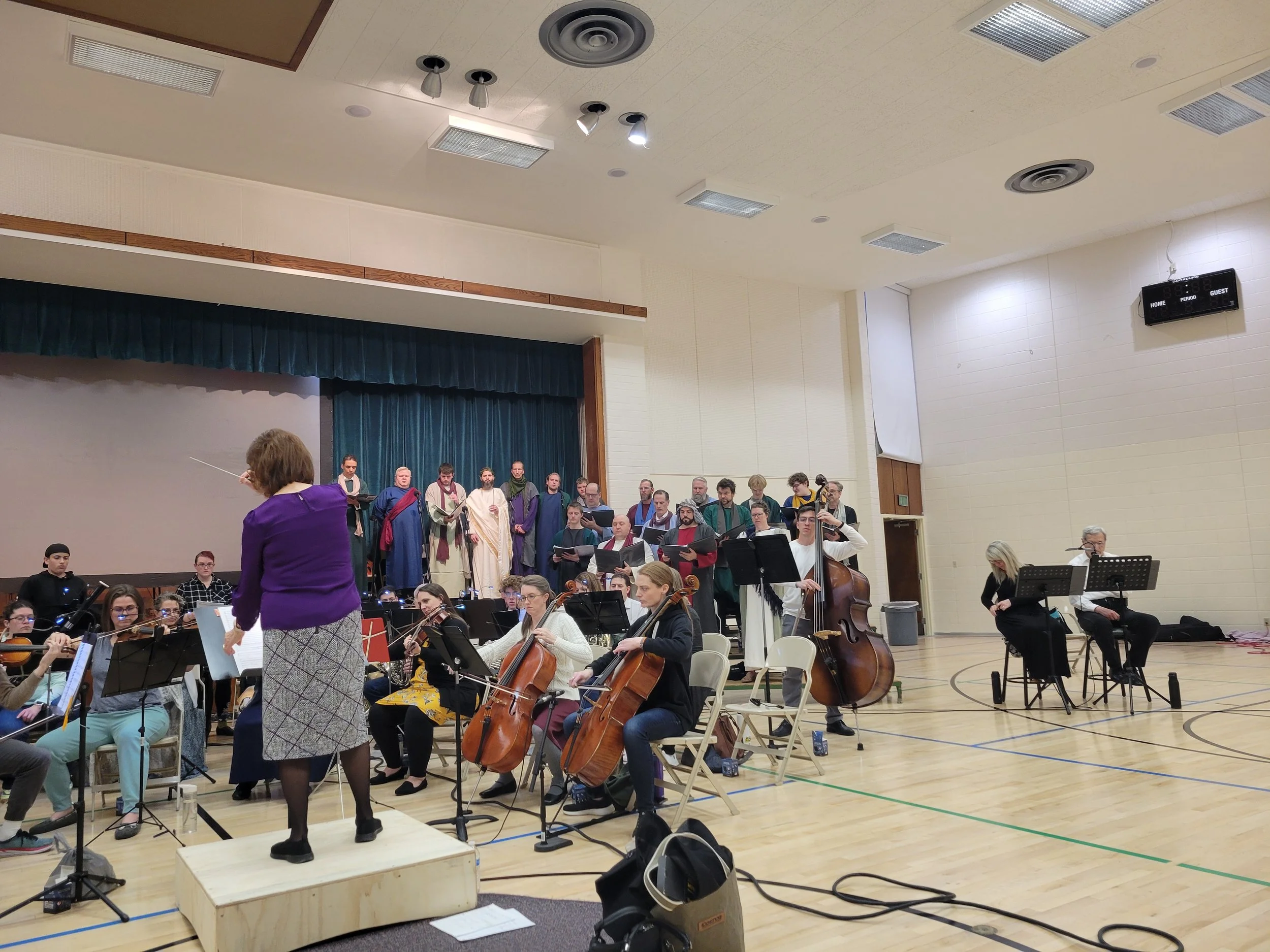 Orchestra rehearsal in a gymnasium, with a choir group standing on stage and musicians playing instruments.