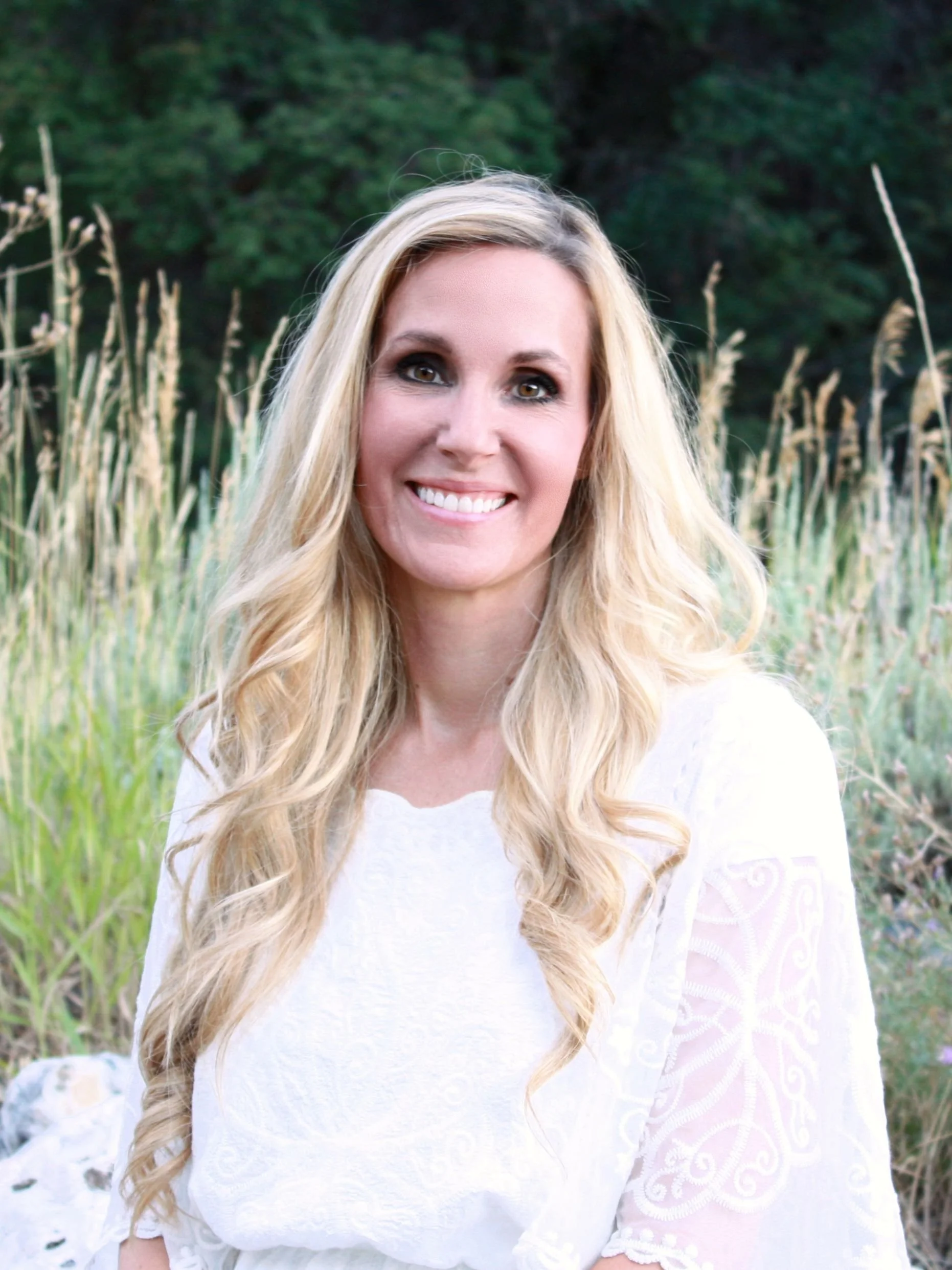 A woman with long blonde hair smiling outdoors in a field with tall grass and trees in the background.