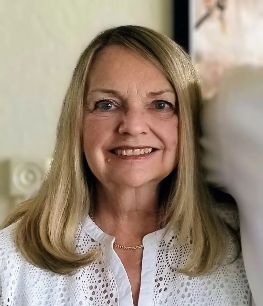 A middle-aged woman with shoulder-length blonde hair, blue eyes, and a bright smile, wearing a white embroidered blouse and a gold chain necklace, indoors against a light-colored wall.