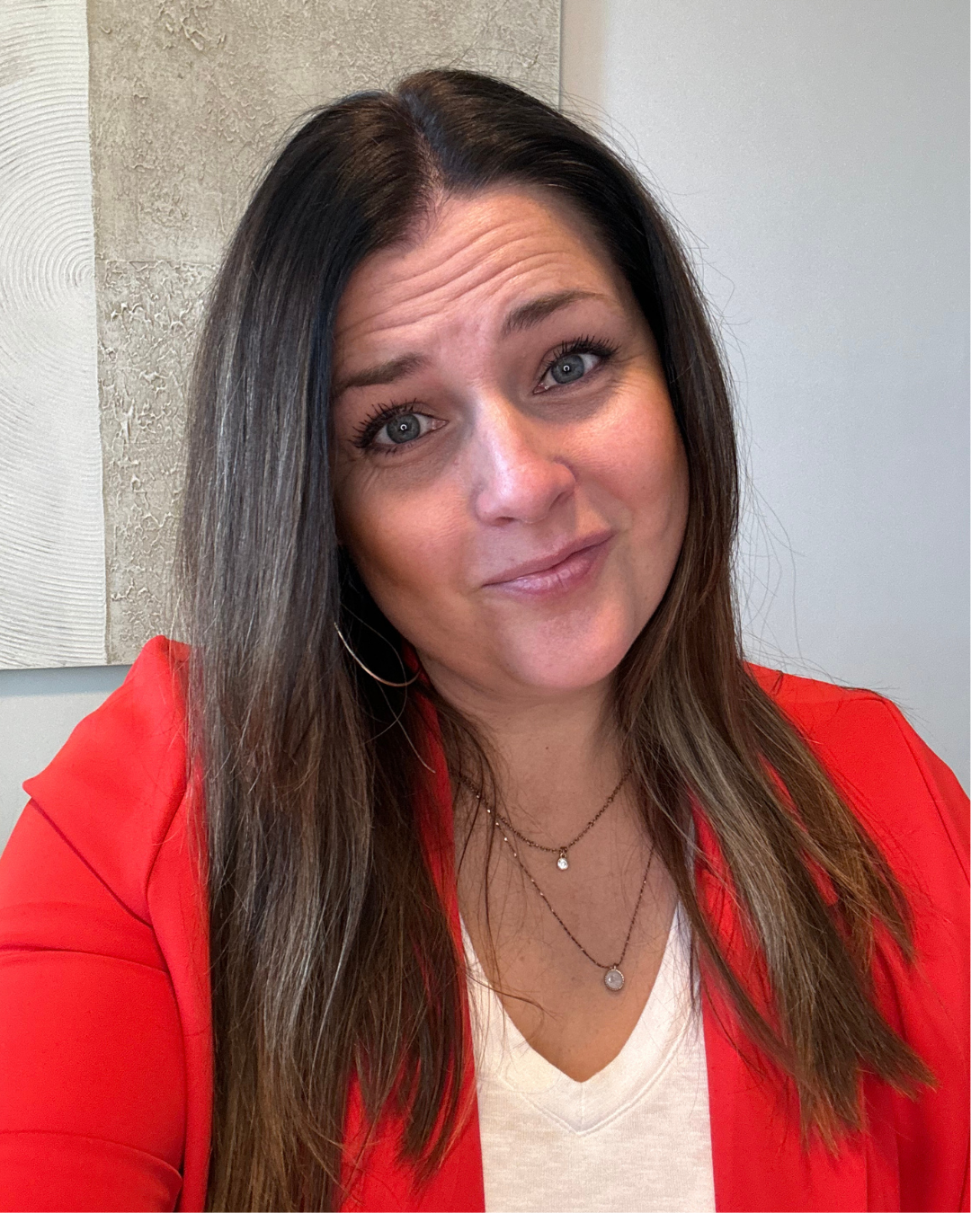 A woman with long brown hair and blue eyes, wearing a red blazer and white top, smiling slightly at the camera, with layered necklaces and hoop earrings, in an indoor setting with textured and smooth wall backgrounds.