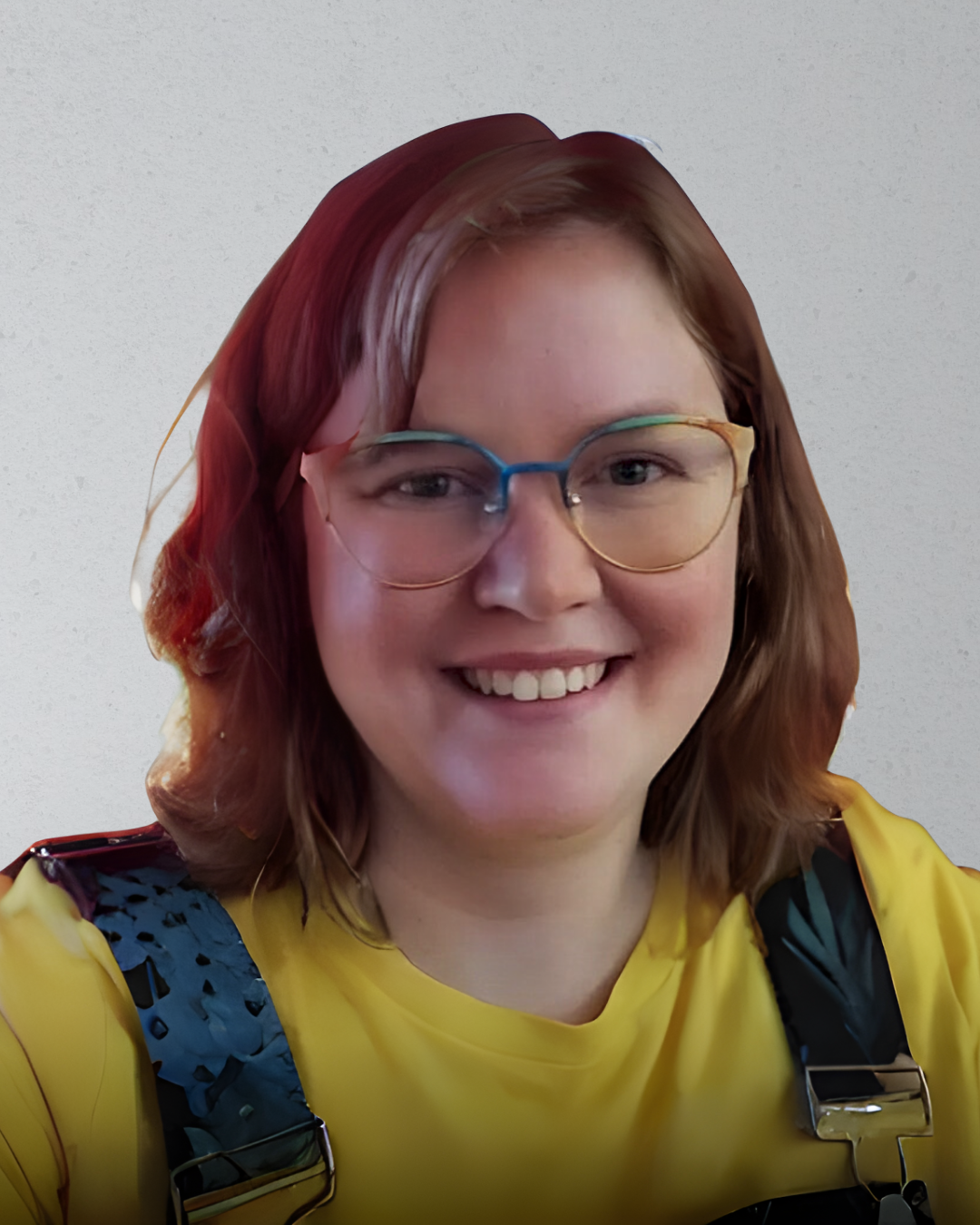 Close-up of a smiling woman with shoulder-length red hair, wearing rainbow-colored glasses and a yellow shirt, standing against a plain white wall.