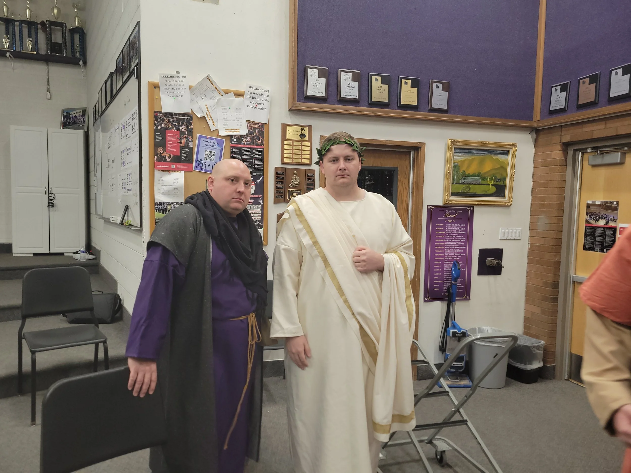 Two men are standing indoors in front of a bulletin board, one dressed as a Roman or Greek figure in white robes with a laurel wreath, and the other in dark robes with a cloak. The room has chairs, posters, and framed awards on the walls.