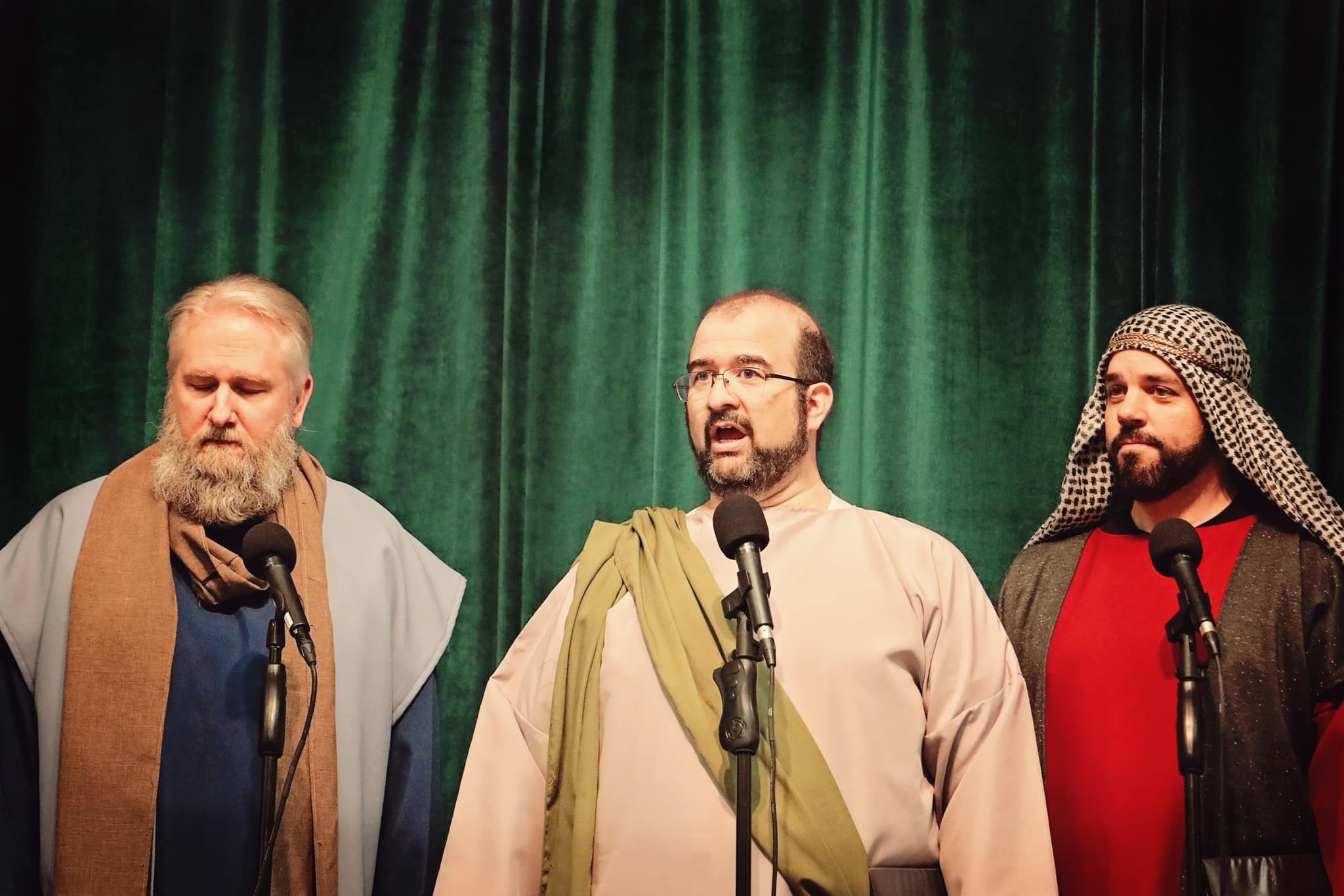 Three men wearing biblical costumes stand in front of a green curtain, each speaking into a microphone during a stage performance.
