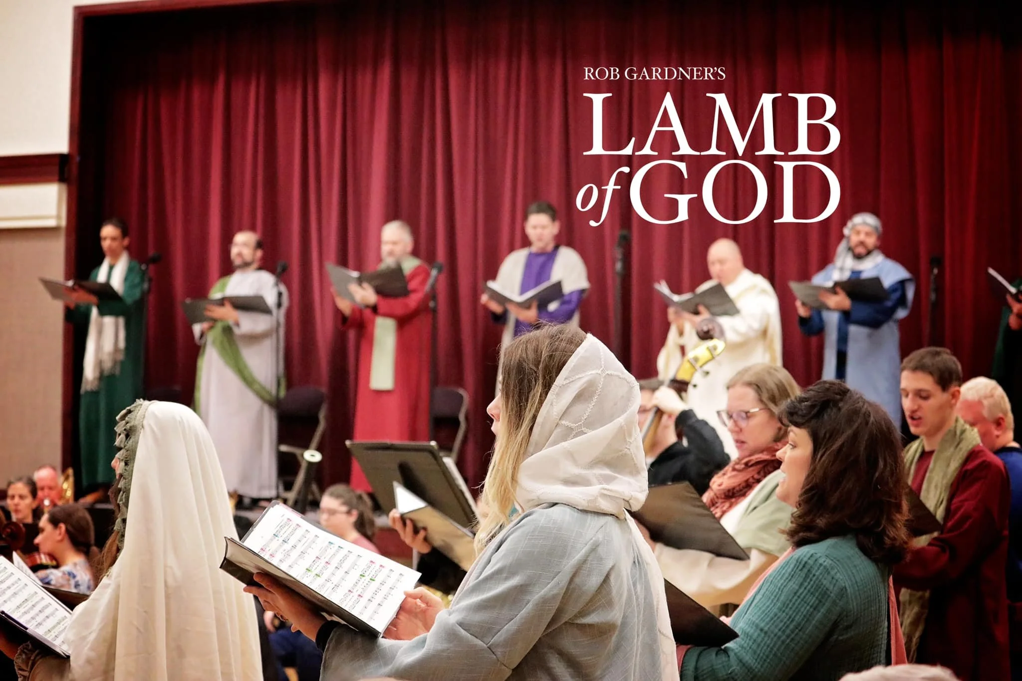 People participating in a choir rehearsal for 'Lamb of God' play on stage with a red curtain backdrop.