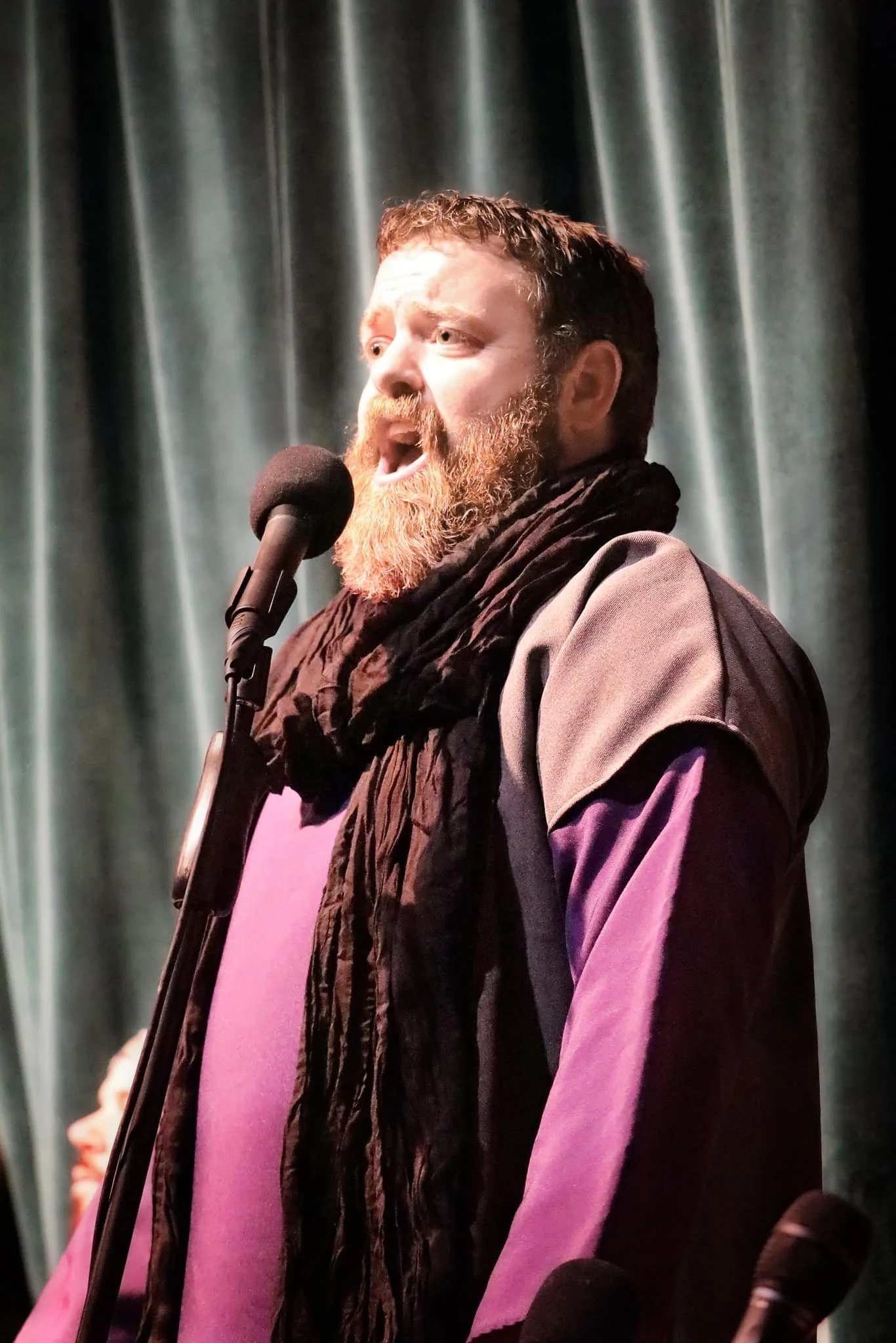 A man with a beard and short hair singing or speaking into a microphone on stage with curtains in the background.
