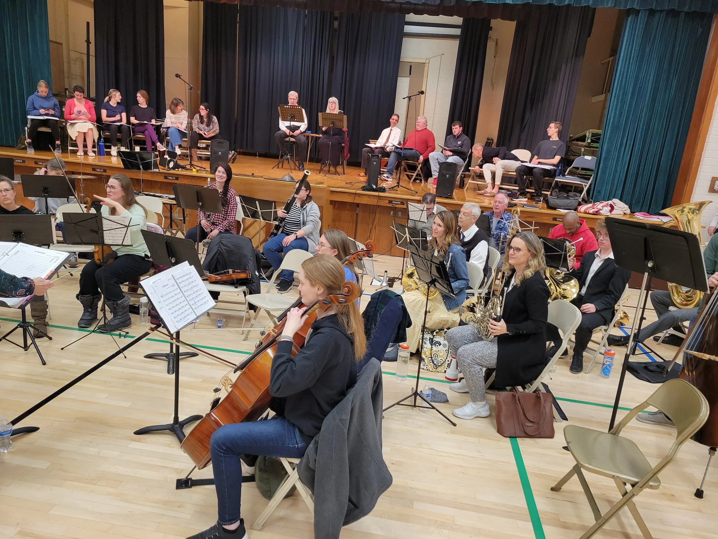 An orchestra and choir rehearsal in a gymnasium with musicians playing string, woodwind, and brass instruments, and choir members sitting on a stage with music stands, black curtains, and microphones.