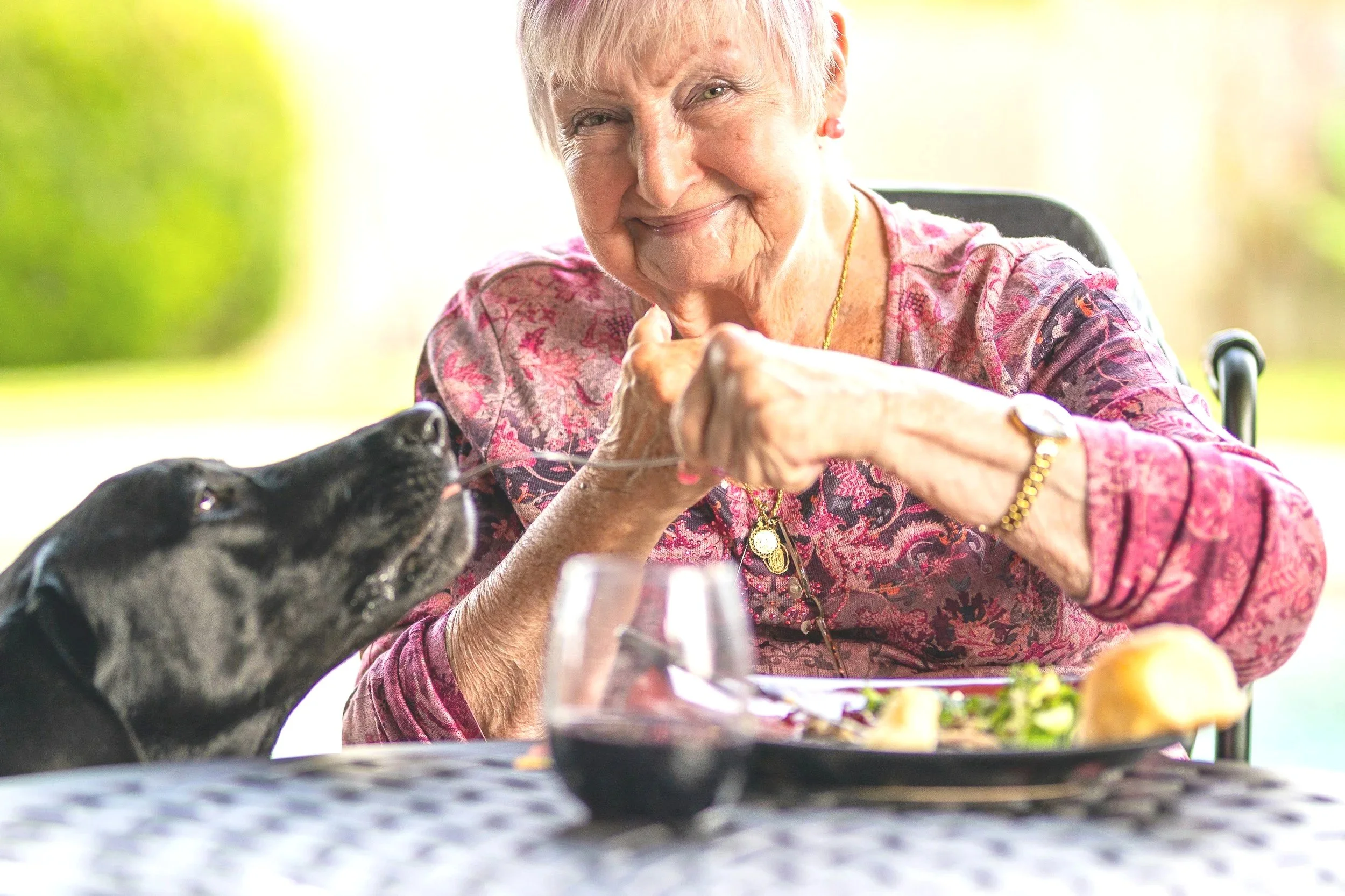 An elderly woman with a big smile sitting at a table outdoors, holding a dog’s paw, with a glass of red wine and a plate of food on the table.