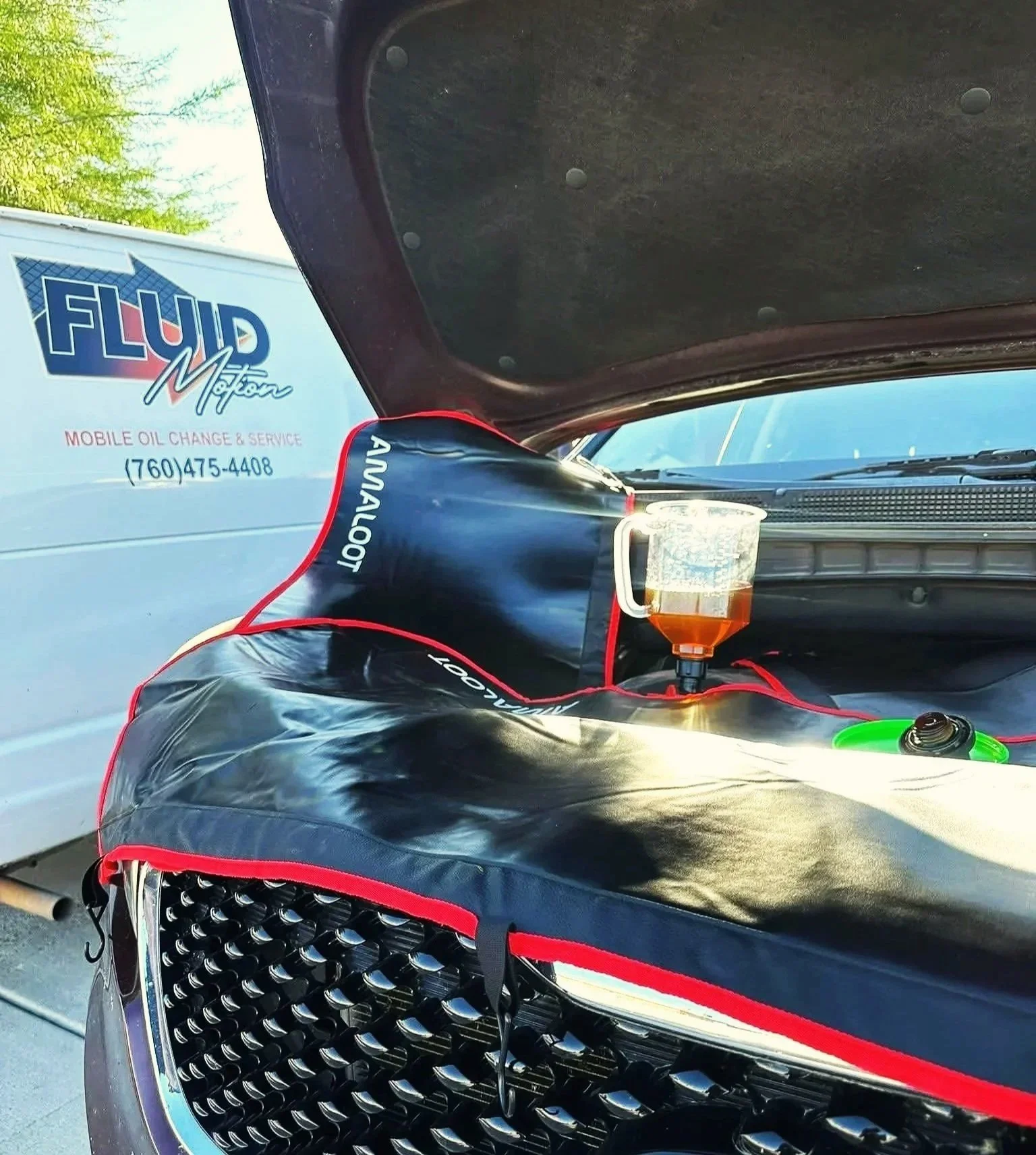 Car hood with a black maintenance mat labeled 'DIPSTICK, OIL, COOLANT' in red, a funnel filled with dark oil, and a green tool on the engine compartment. A white truck with 'Fluid Motion Mobile Oil Change & Service' is visible in the background.