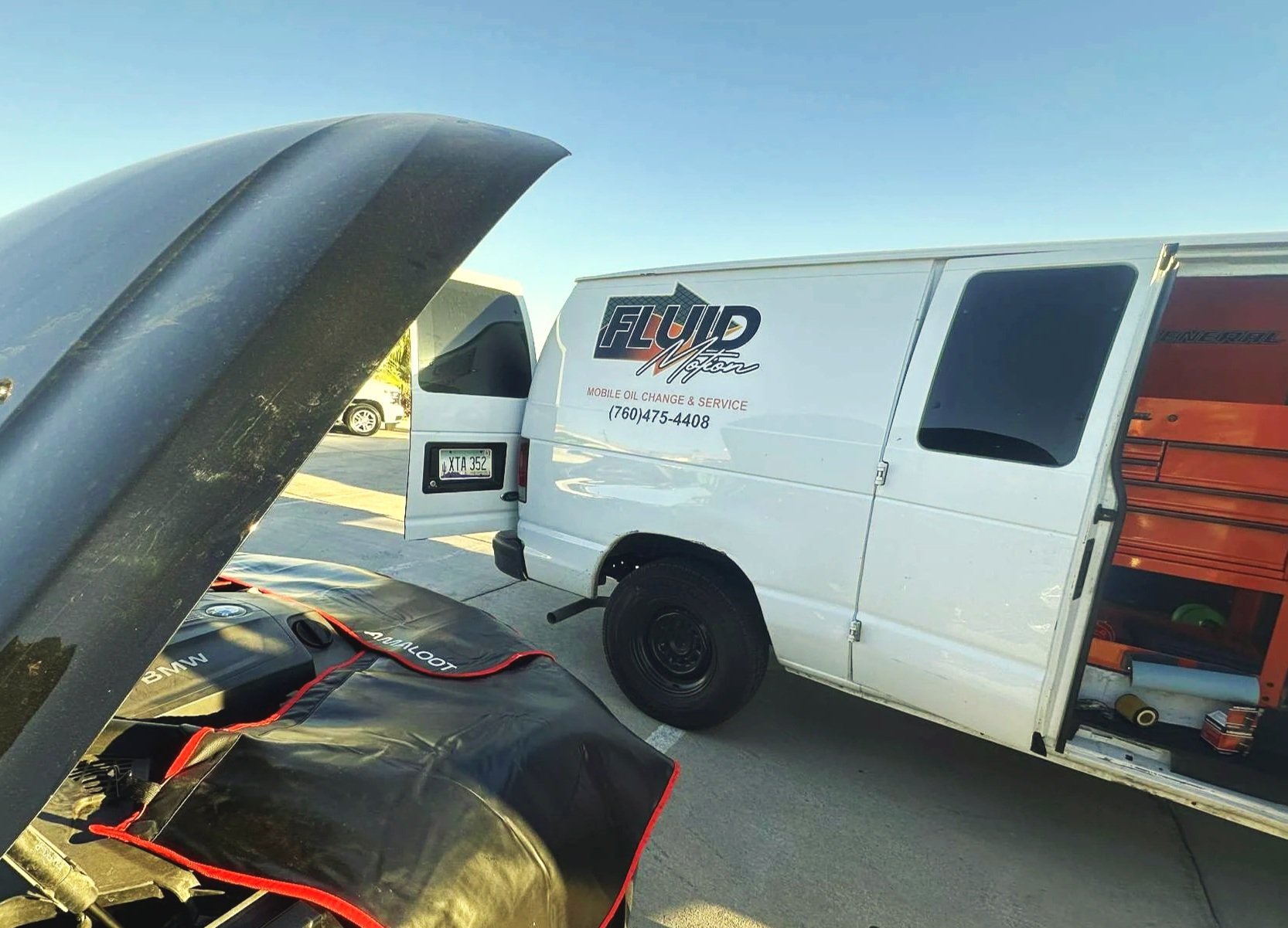 A white service van with orange and black lettering advertising liquid motor oil change and service, parked in a lot with its side door open revealing tools inside. In the foreground, part of a car with an open hood and engine cover is visible.
