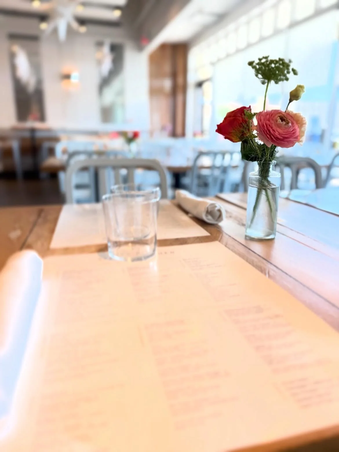 A restaurant table set with a menu, a glass of water, a rolled napkin, and a small vase with pink and red flowers near the window.