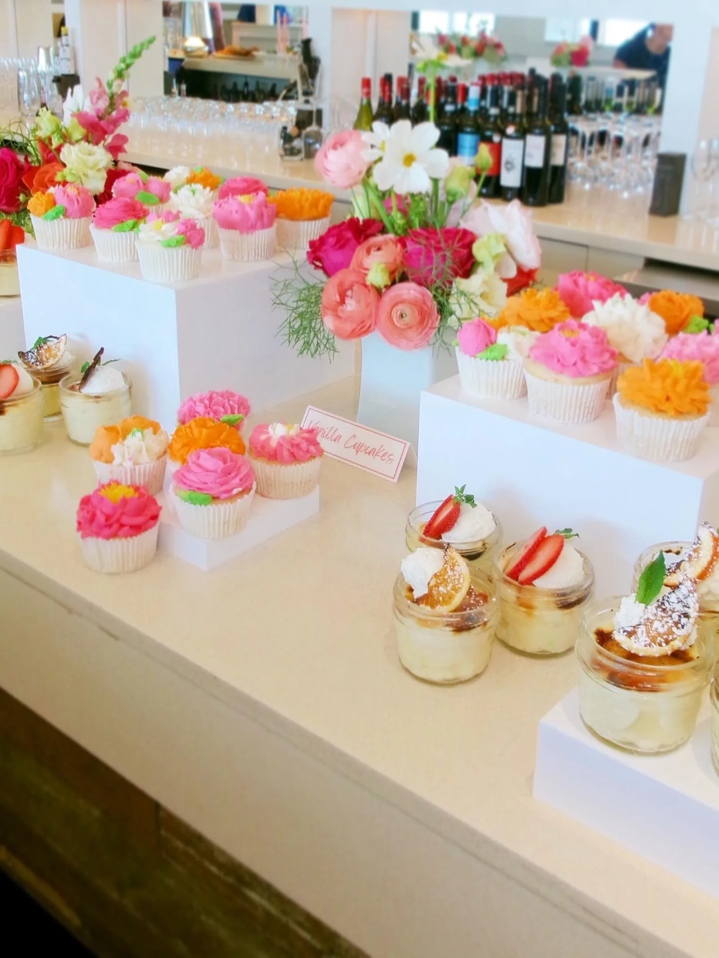 Display of colorful cupcakes with pink, orange, and white frosting, along with strawberry-topped parfaits and a floral arrangement of pink, white, and green flowers on a table at a party or café.