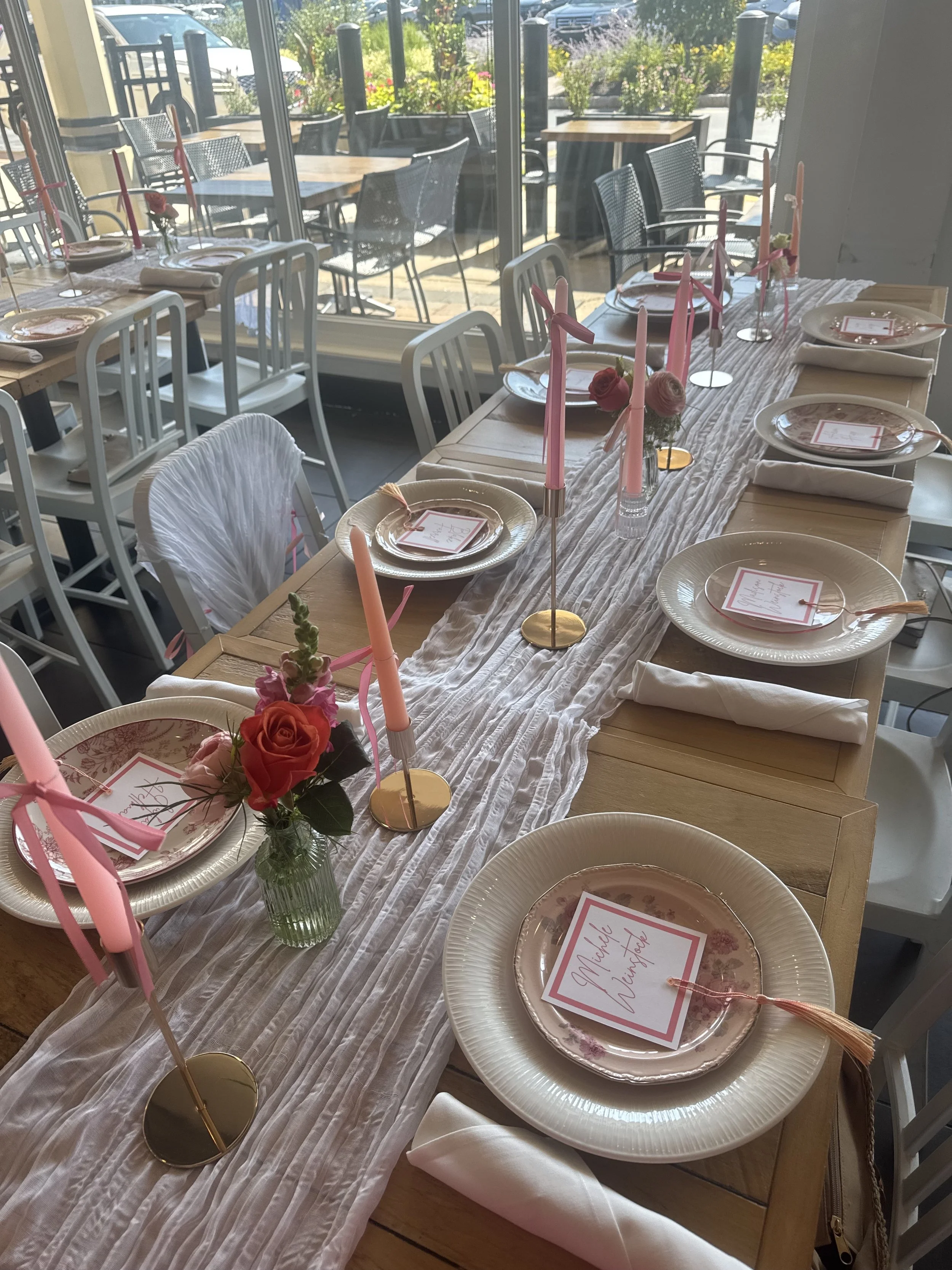 A long wooden dining table set for a celebration with pink candles, small flower arrangements, pink and cream plates, and place cards that read 'Michał Wiśniewski.' The table has a white table runner and white cloth napkins, with chairs around it. In
