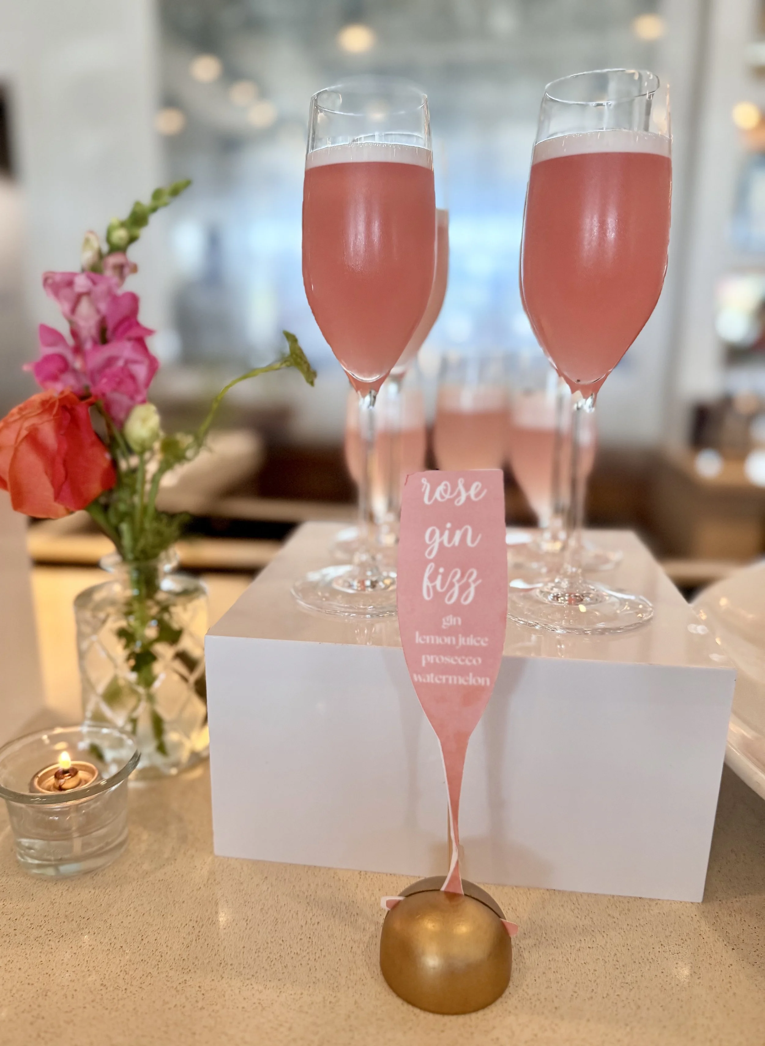 Two glasses of pink rosé gin fizz cocktails on a white box, with a small flower arrangement and a lit candle nearby, and a pink tag listing ingredients including gin, lemon juice, prosecco, and watermelon.