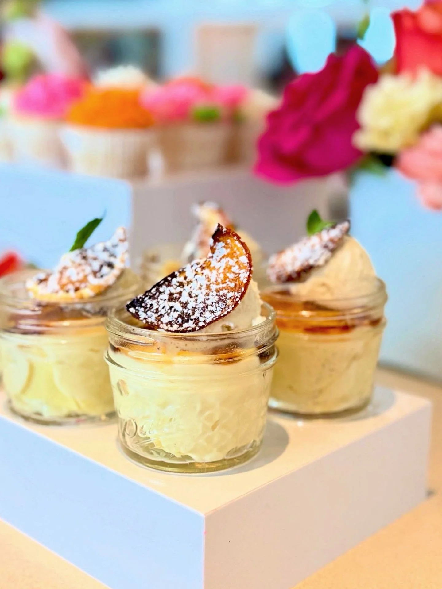 Three glass jars of creamy dessert topped with a dusting of powdered sugar and a small slice of dried orange, each garnished with a small mint leaf, displayed on a white cake stand with blurred pink and red flowers in the background.