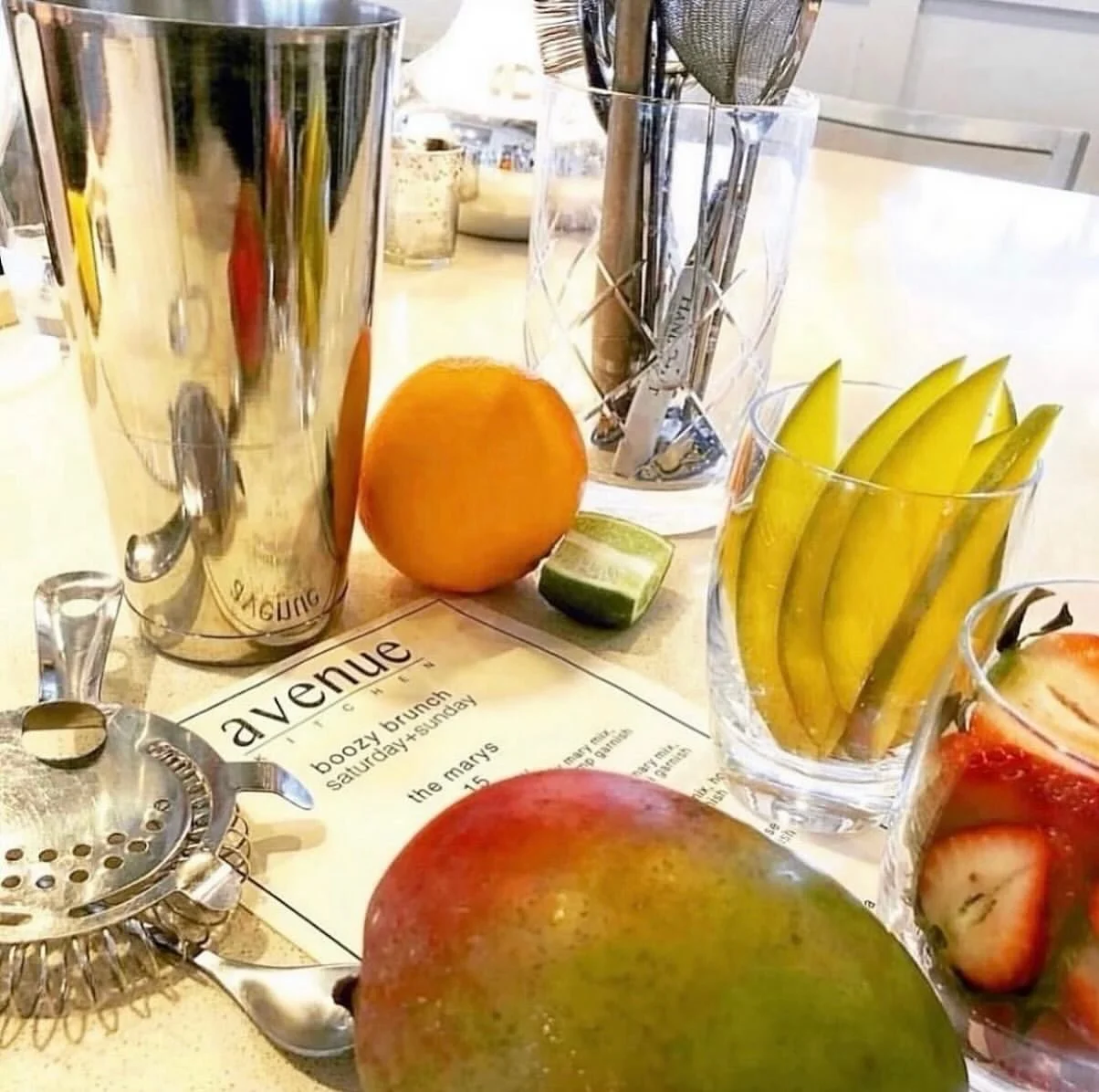 A variety of fresh fruits including an apple, banana, strawberry, orange, and lime on a table, with cocktail shakers, glasses, and a menu in the background.