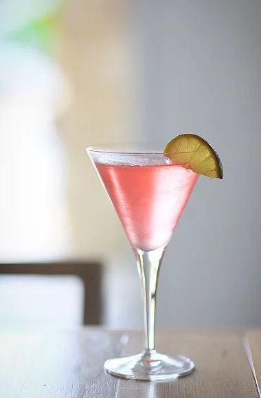 A pink cocktail garnished with a lime wedge in a martini glass on a table.