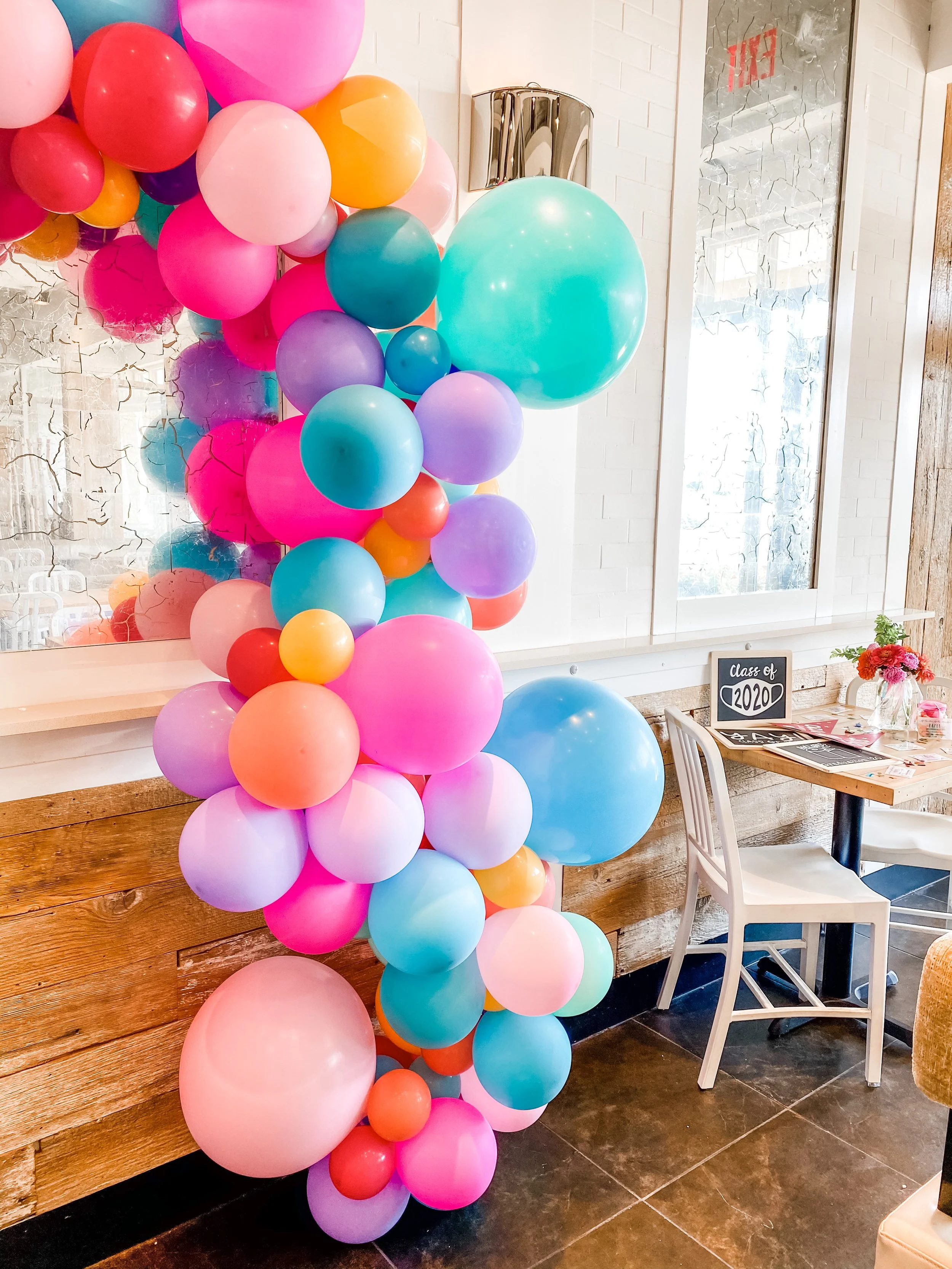 A colorful balloon display with pink, purple, blue, orange, and red balloons arranged indoors next to a wooden wall, cafe table, and window.