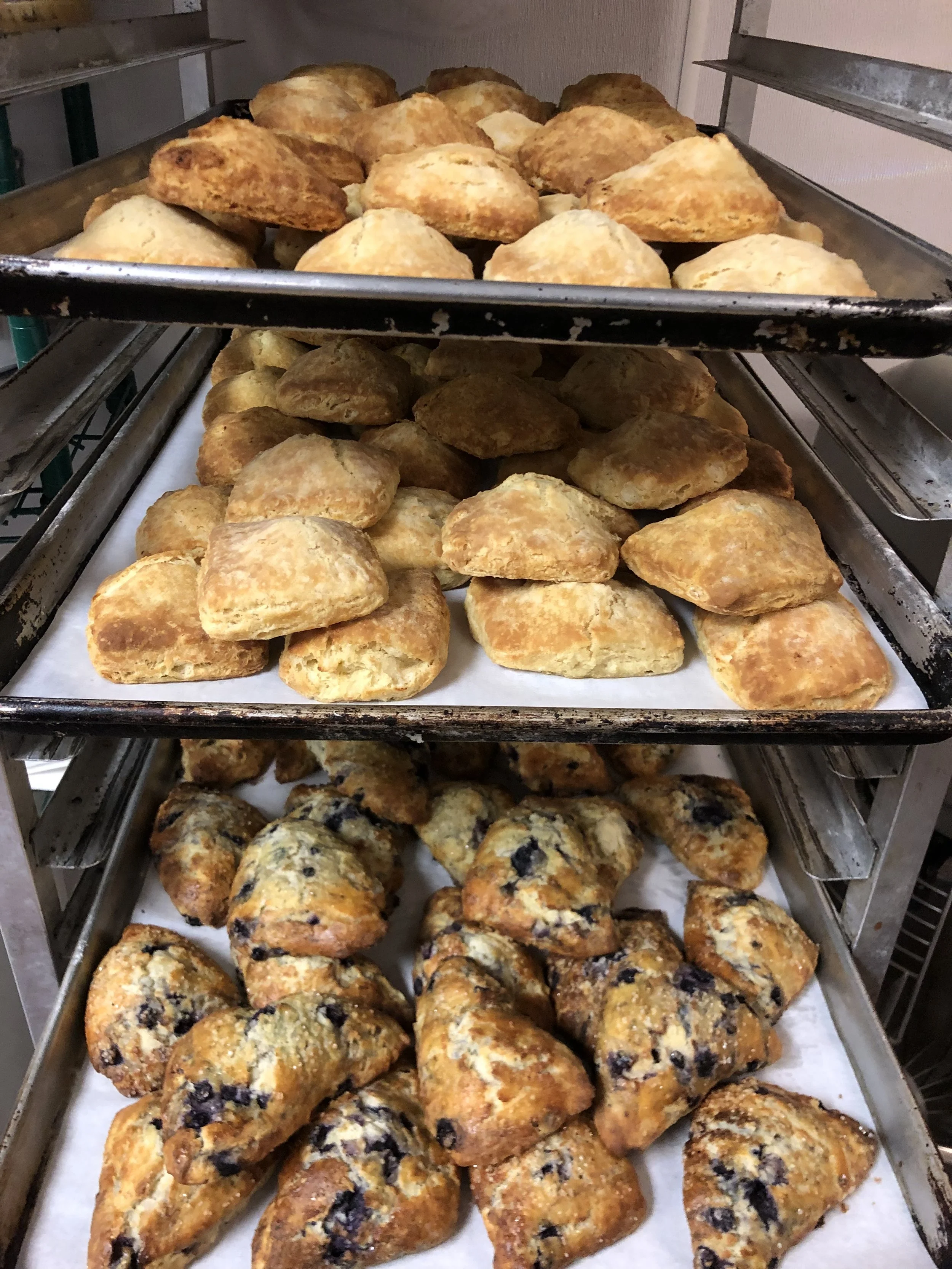 Three shelves with baked goods: top shelf has scones, middle shelf has bread rolls, bottom shelf has blueberry muffins on parchment paper.