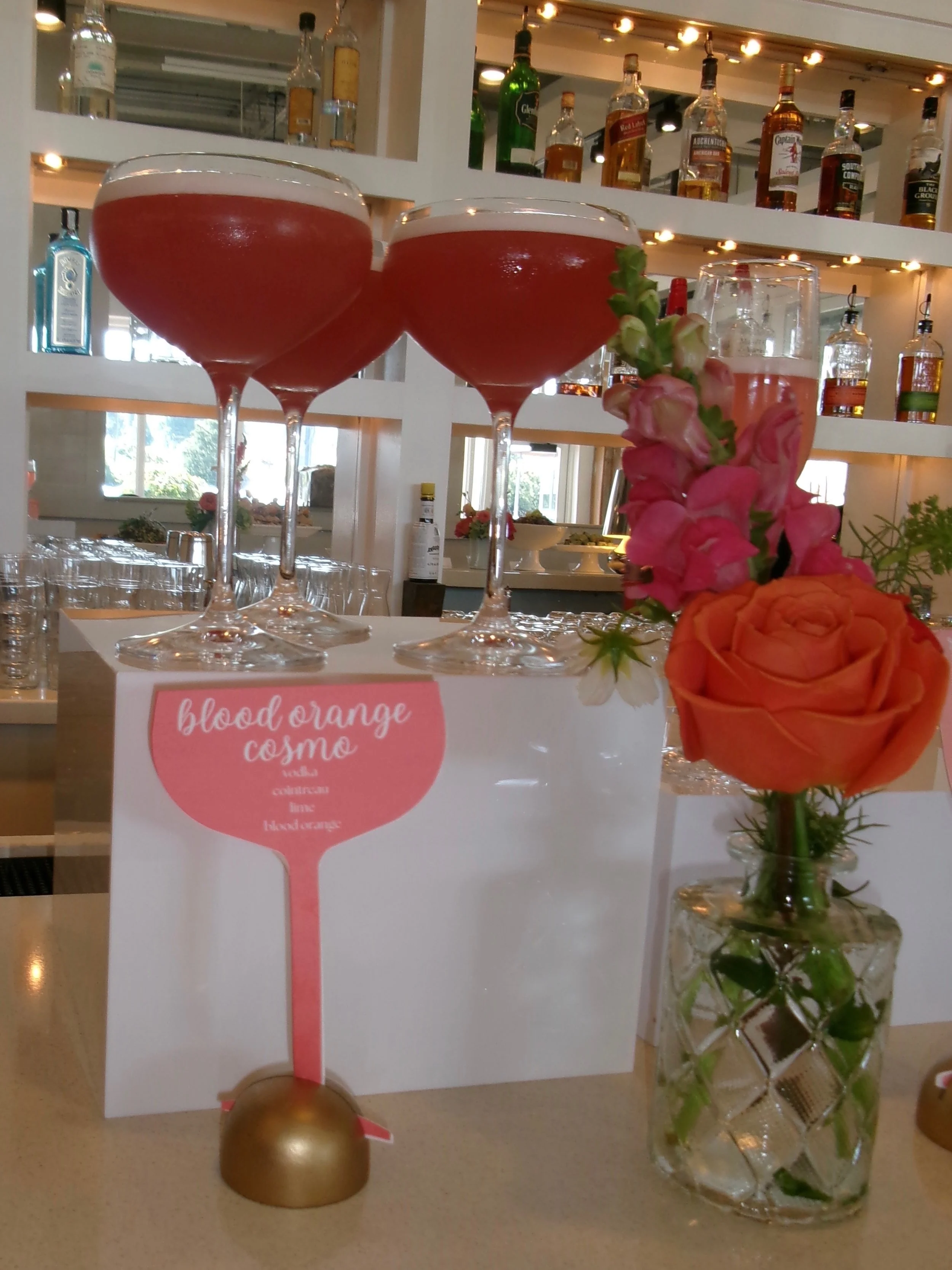 Two cocktails in stemmed glasses with pink liquid, a glass with pink and peach flowers, and a sign that says 'blood orange cosme' in a bar or restaurant setting.