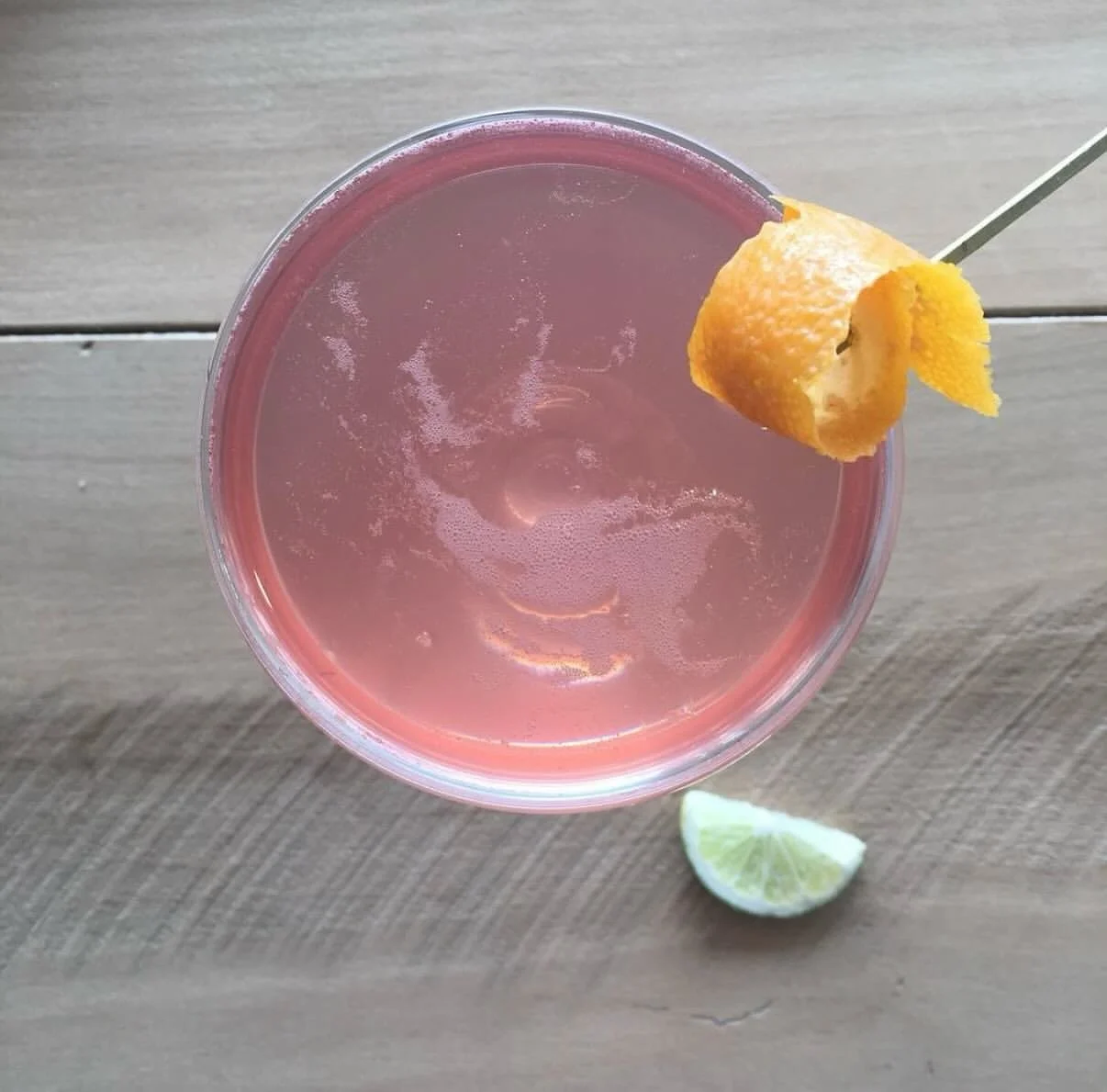 A pink cocktail garnished with lemon and lime wedges on a wooden table.