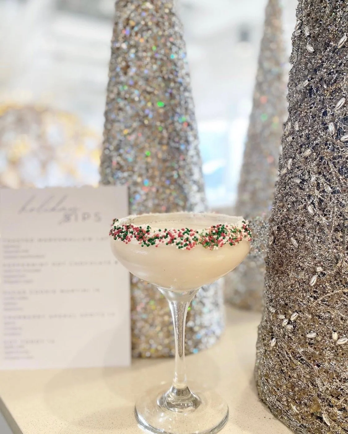 A cocktail glass with a white drink, decorated with red and green sprinkles around the rim, on a table with glittering decorated cone-shaped objects in the background.