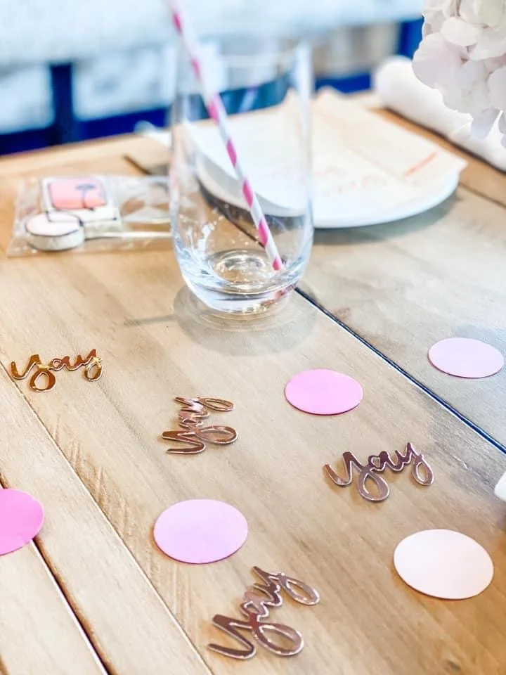 Decorated wooden table with pink paper circles, rose gold 'you' and 'me' cutouts, empty glass with red and white striped straw, and a white plate in the background, likely for a celebration or romantic event.