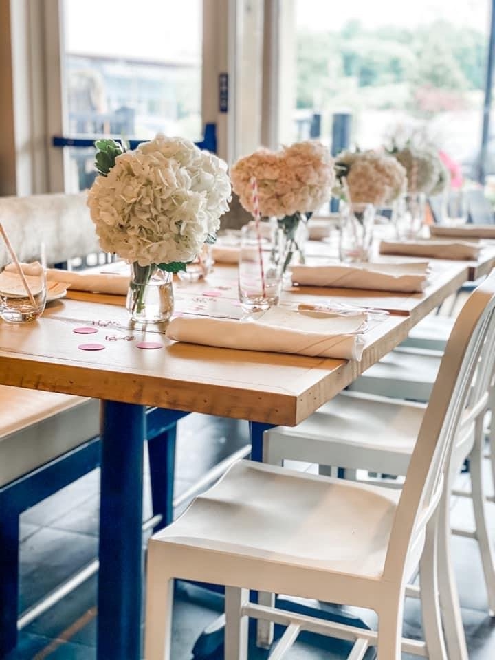 A wooden dining table set for a celebration with white hydrangea flower centerpieces in glass vases, beige napkins, and pink confetti on the tablecloth, in a bright room with large windows.