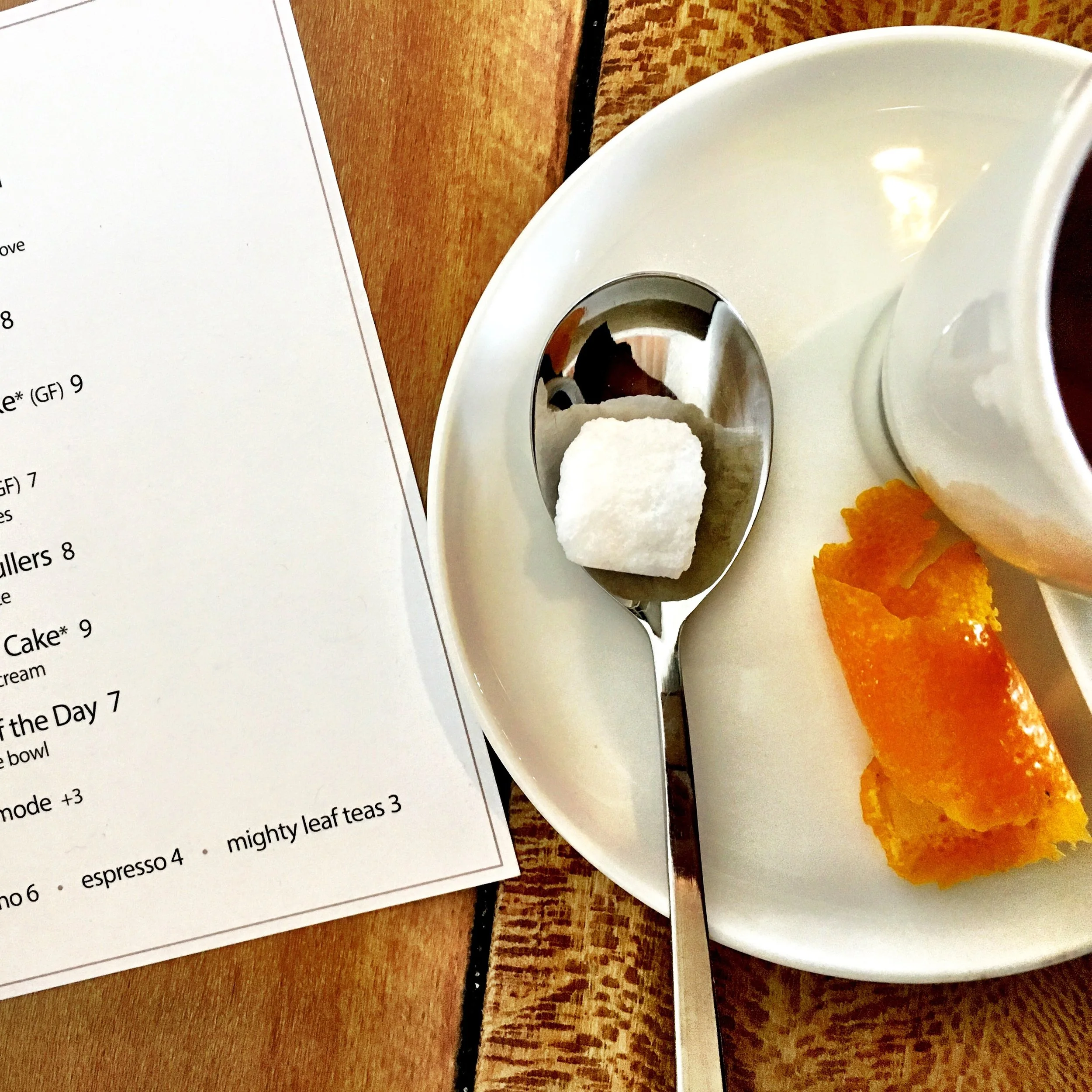 A white plate holding a spoon with a sugar cube, a piece of orange peel, and a white cup with a drink. Part of a menu is visible on the left side of the image.