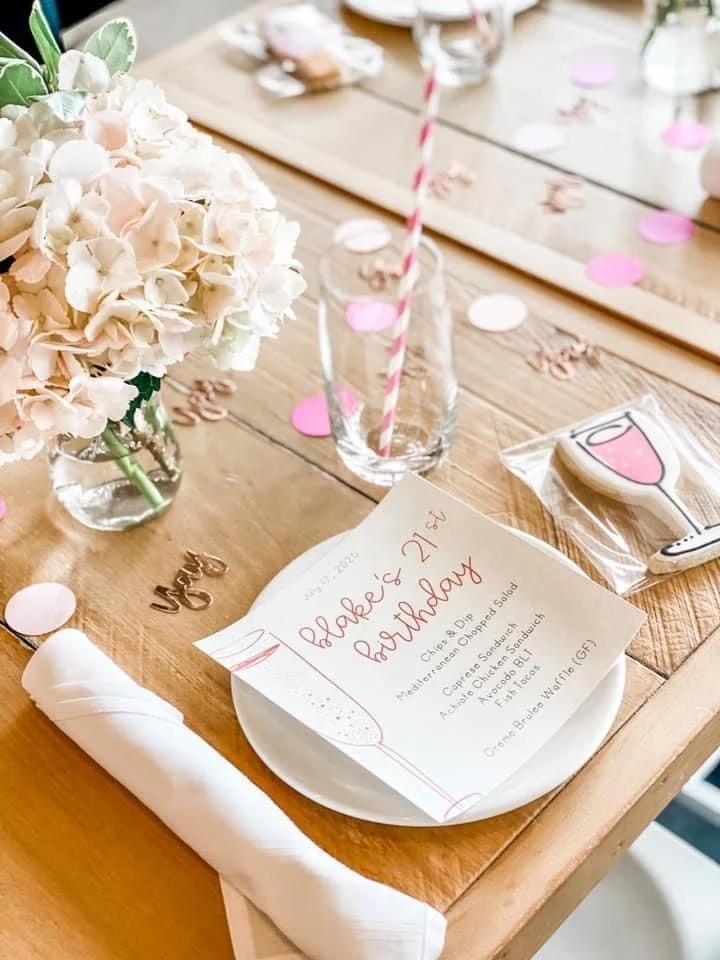 Table setting for a birthday celebration with a pink theme. Includes a menu card, pink confetti, a glass with a pink striped straw, a floral centerpiece with light pink hydrangeas, and a decorated napkin with a party blowout in a birthday hat.