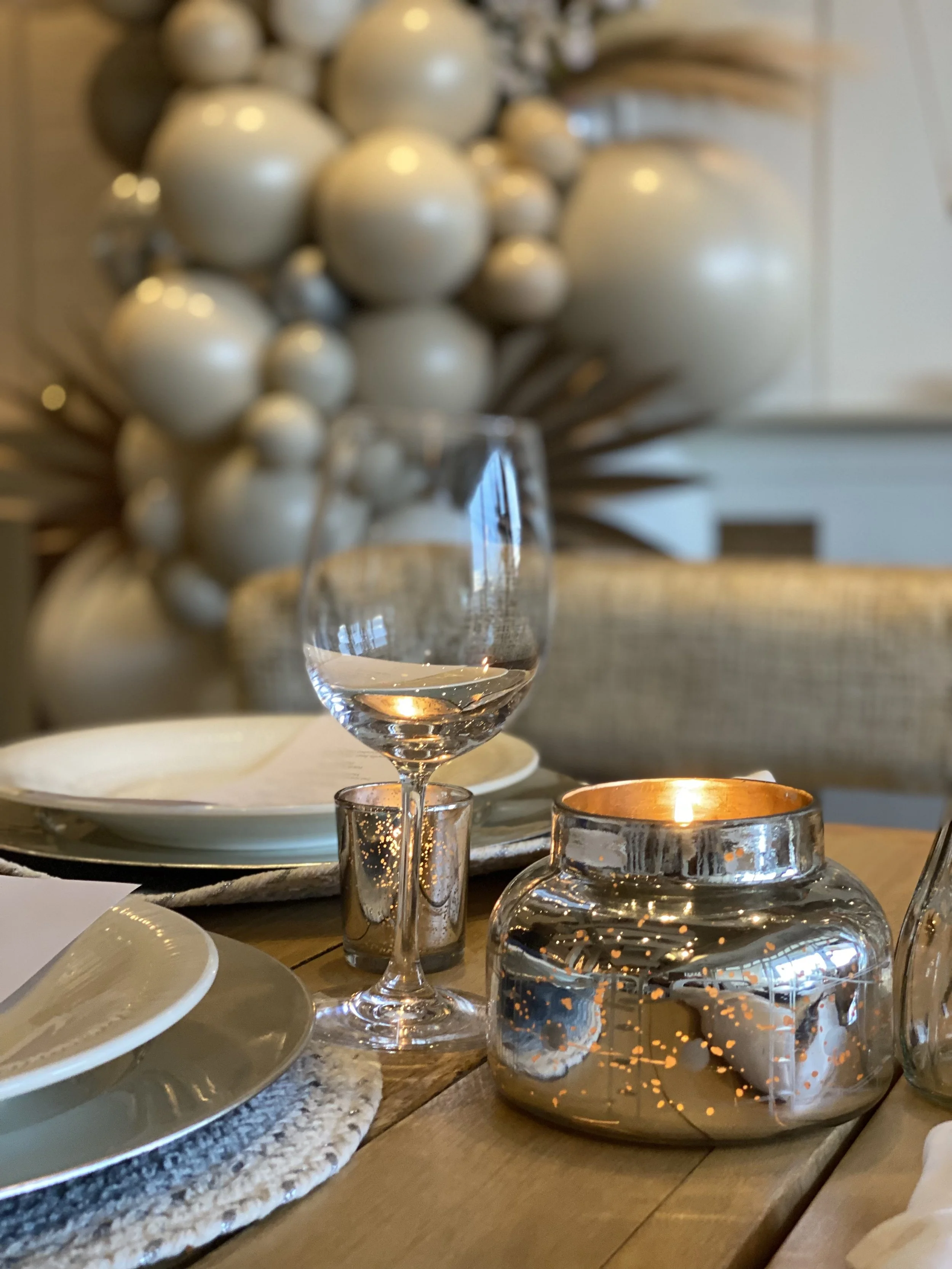 Elegant table setting with a wine glass, a lit candle in a metallic holder, and plates on a woven placemat. In the background, decorative white ornaments resembling balloons or bubbles arranged on a wall.