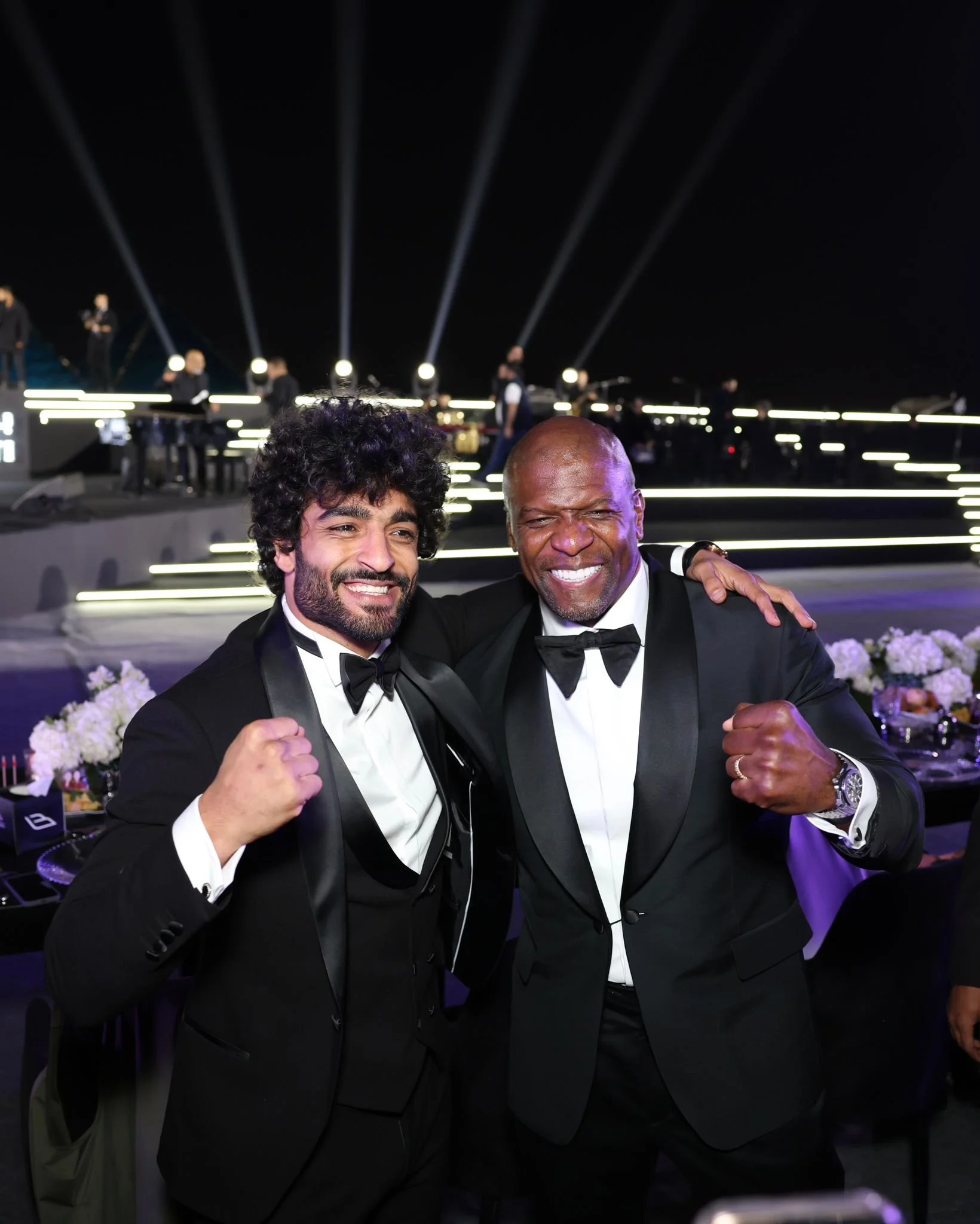Two men in tuxedos pose together with fists raised, smiling at a formal event with a stage and performers in the background.