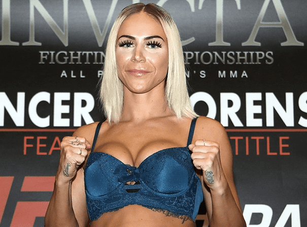 Female fighter with platinum blonde hair posing with clenched fists in front of a black and red promotional background, wearing a blue lace bra.