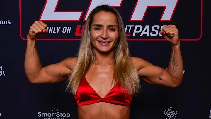 A woman with long blonde hair and a red sports bra posing with her arms flexed in front of a black and red UFC background. She is smiling and showing her muscular arms.