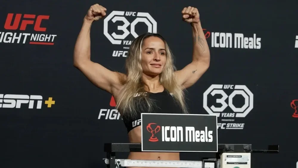 A female mixed martial artist flexing her biceps during a weigh-in, standing in front of a UFC backdrop.