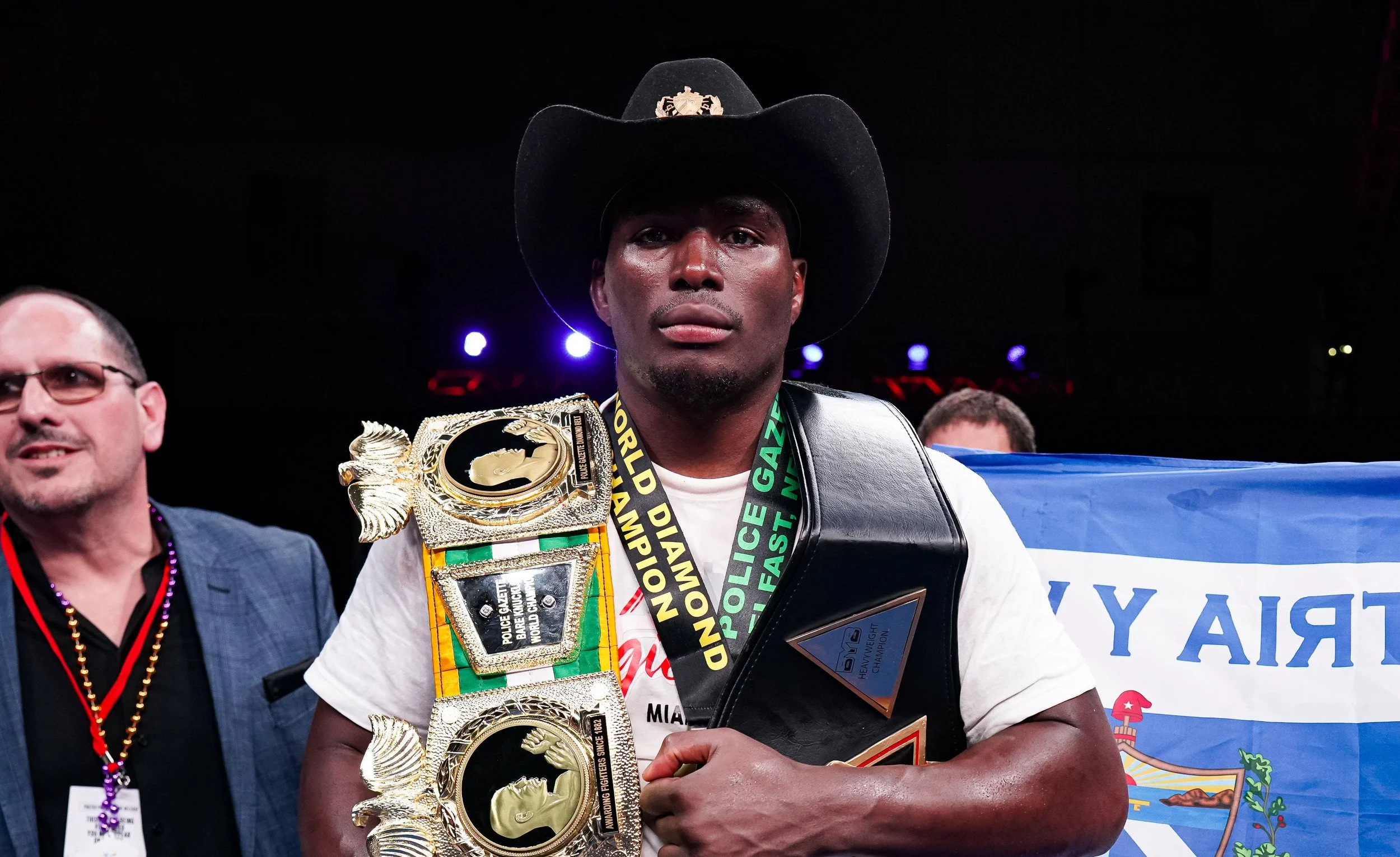 A young man wearing a cowboy hat and holding a large championship belt over his shoulder at a sports event, with a person in glasses and a badge nearby, and a flag with the text 'TRAY Y' in the background.