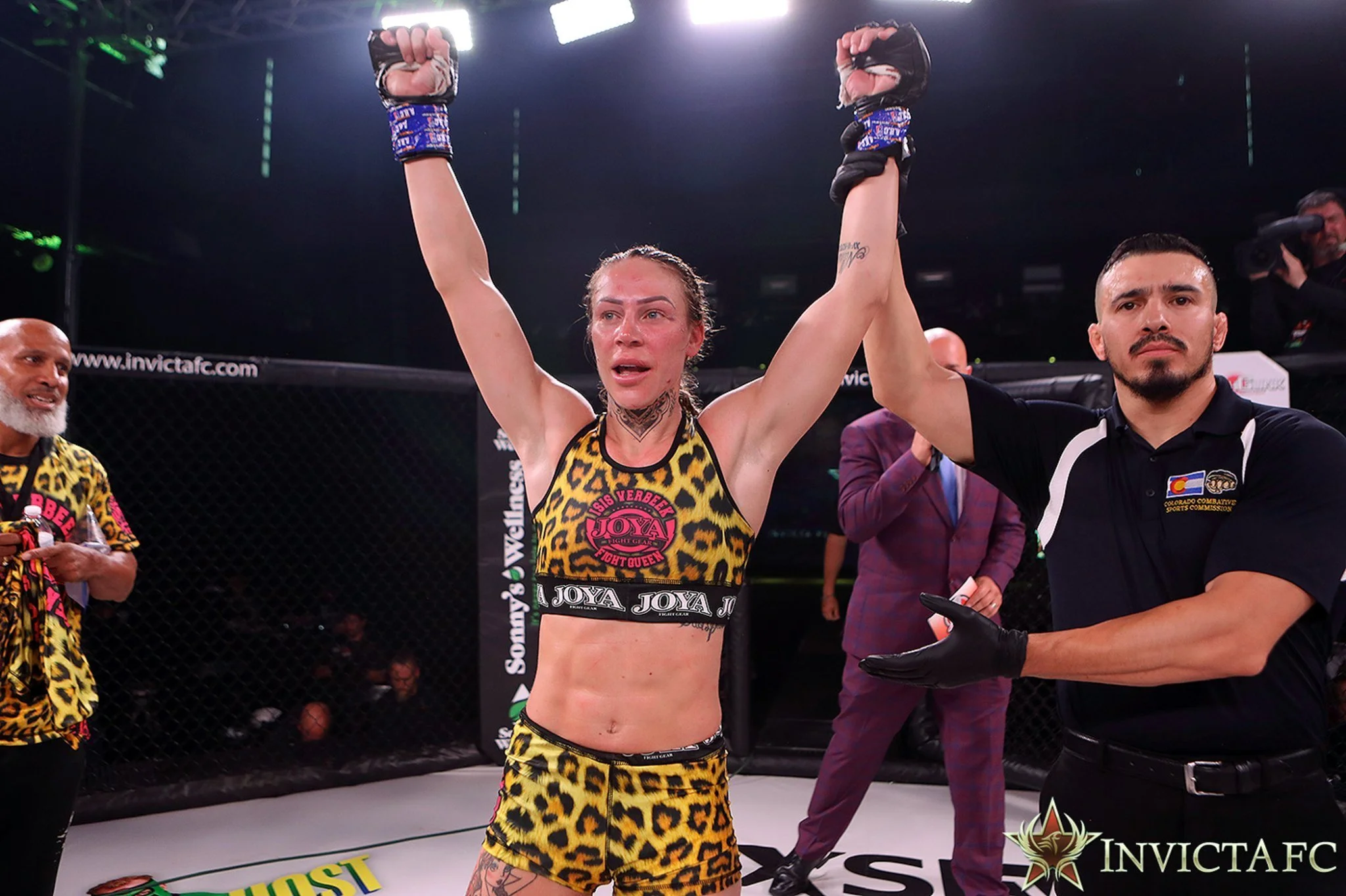 Mixed martial arts fighter in leopard print attire raising her arms in victory inside the octagon, referee holding her wrist, with others in the background celebrating and an 'Invicta FC' logo at the bottom.