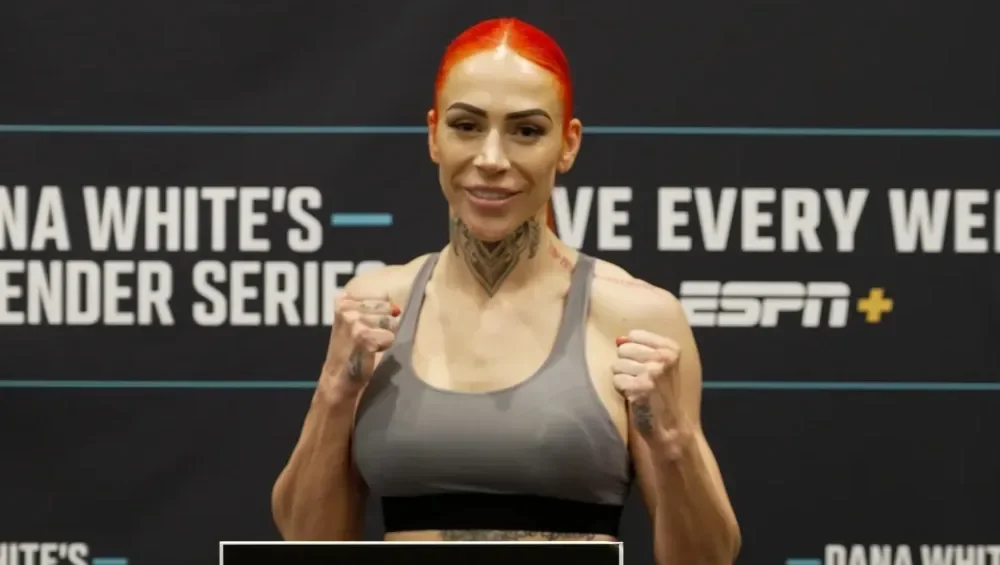 A woman with red hair, visible tattoos, and athletic attire, standing in front of a promotional background for a fighter series on ESPN+. She is holding her fists up in a fighting stance and smiling.
