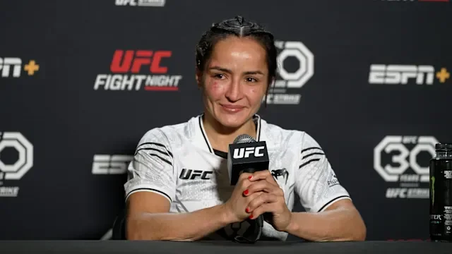 UFC fighter speaking at a press conference, holding a microphone, wearing a white UFC-branded shirt, and looking emotional.
