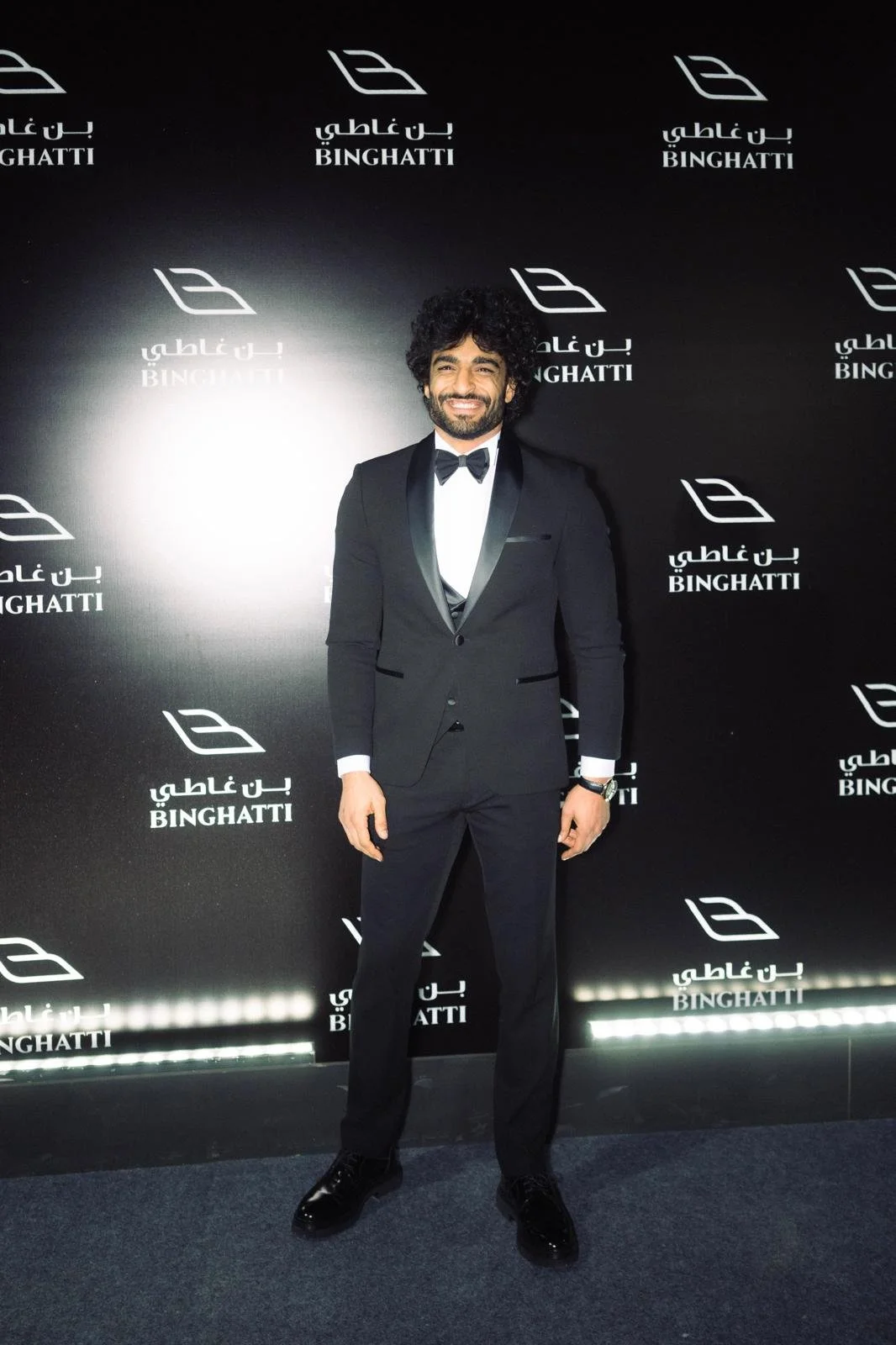 A man in a black tuxedo with a bow tie, standing on a dark carpet in front of a black backdrop with the word "BINGHATTI" and its logo repeatedly printed in white.