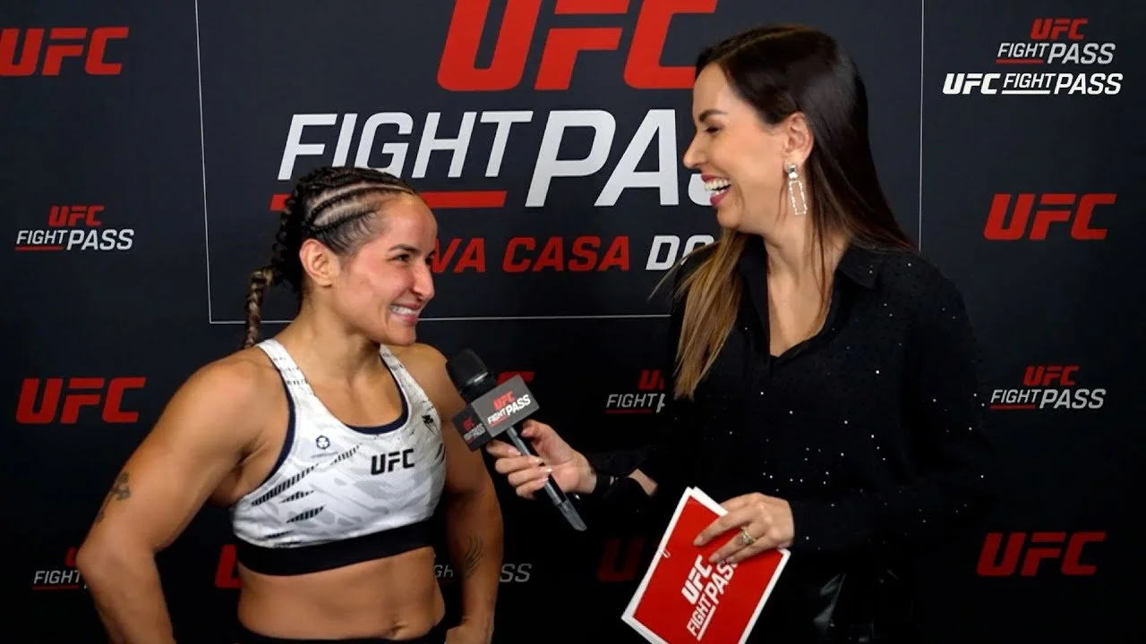 UFC interview scene with a female fighter smiling and talking into a microphone held by a female interviewer, both standing in front of a UFC Fight Pass branded backdrop.