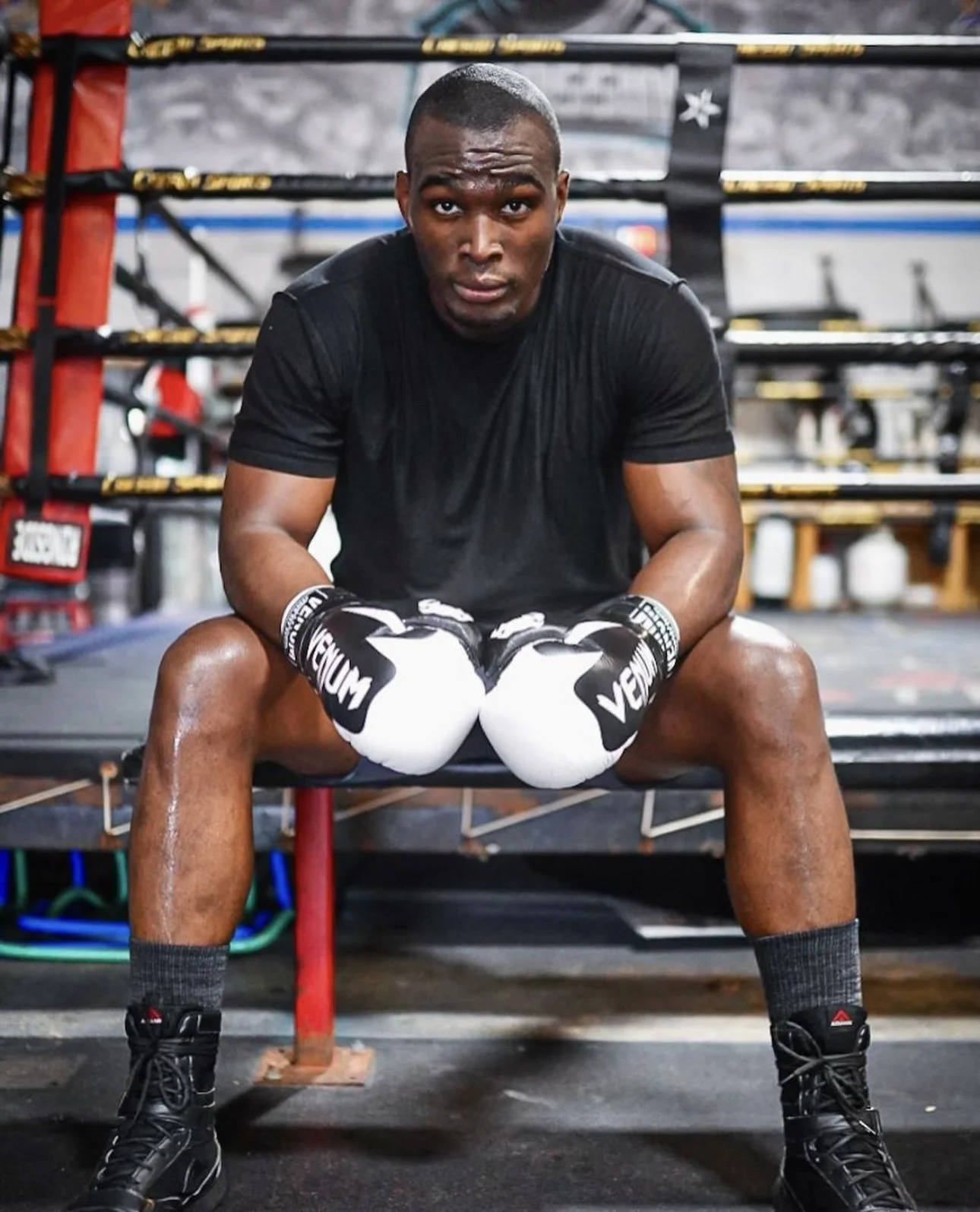 A young man sitting on a boxing bench in a gym, wearing boxing gloves, black shirt, shorts, and boxing shoes, with boxing training equipment in the background.