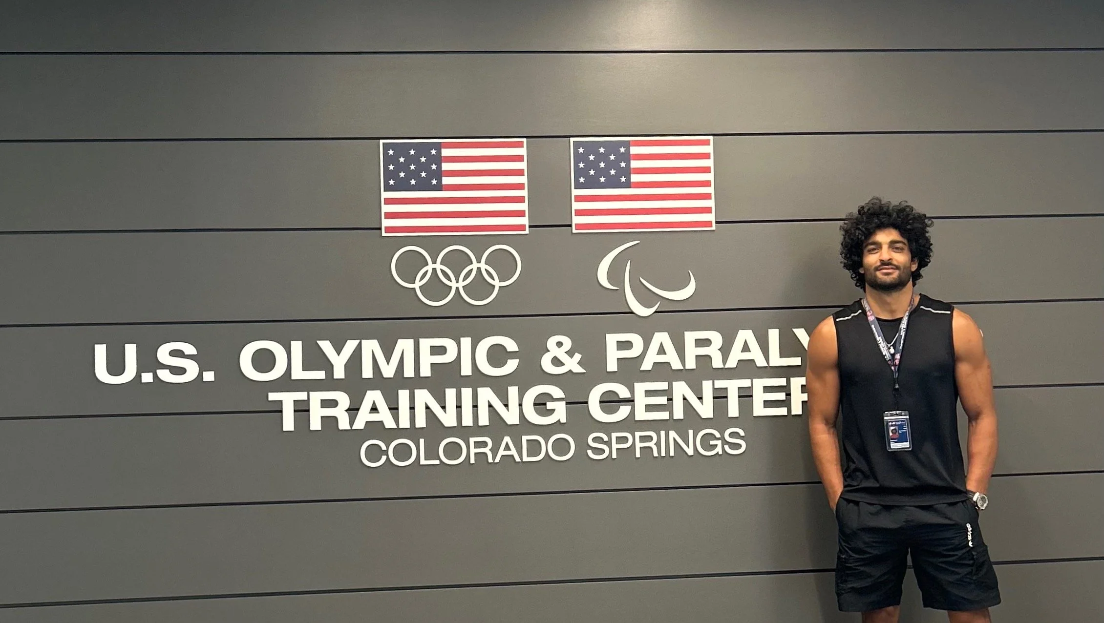 Man standing in front of a sign at the U.S. Olympic & Paralympic Training Center in Colorado Springs with American flags, Olympic rings, and Paralympic symbols.