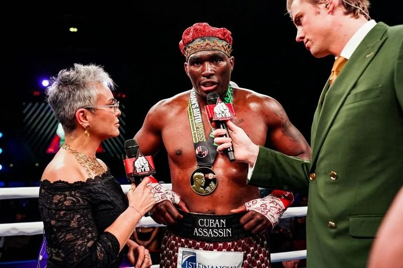 A shirtless male boxer with a red headband being interviewed by two reporters in a boxing ring. The boxer has medals around his neck and a belt that reads 'Cuban Assassin.' One reporter is a woman with short gray hair and glasses, and the other is a man in a green suit.