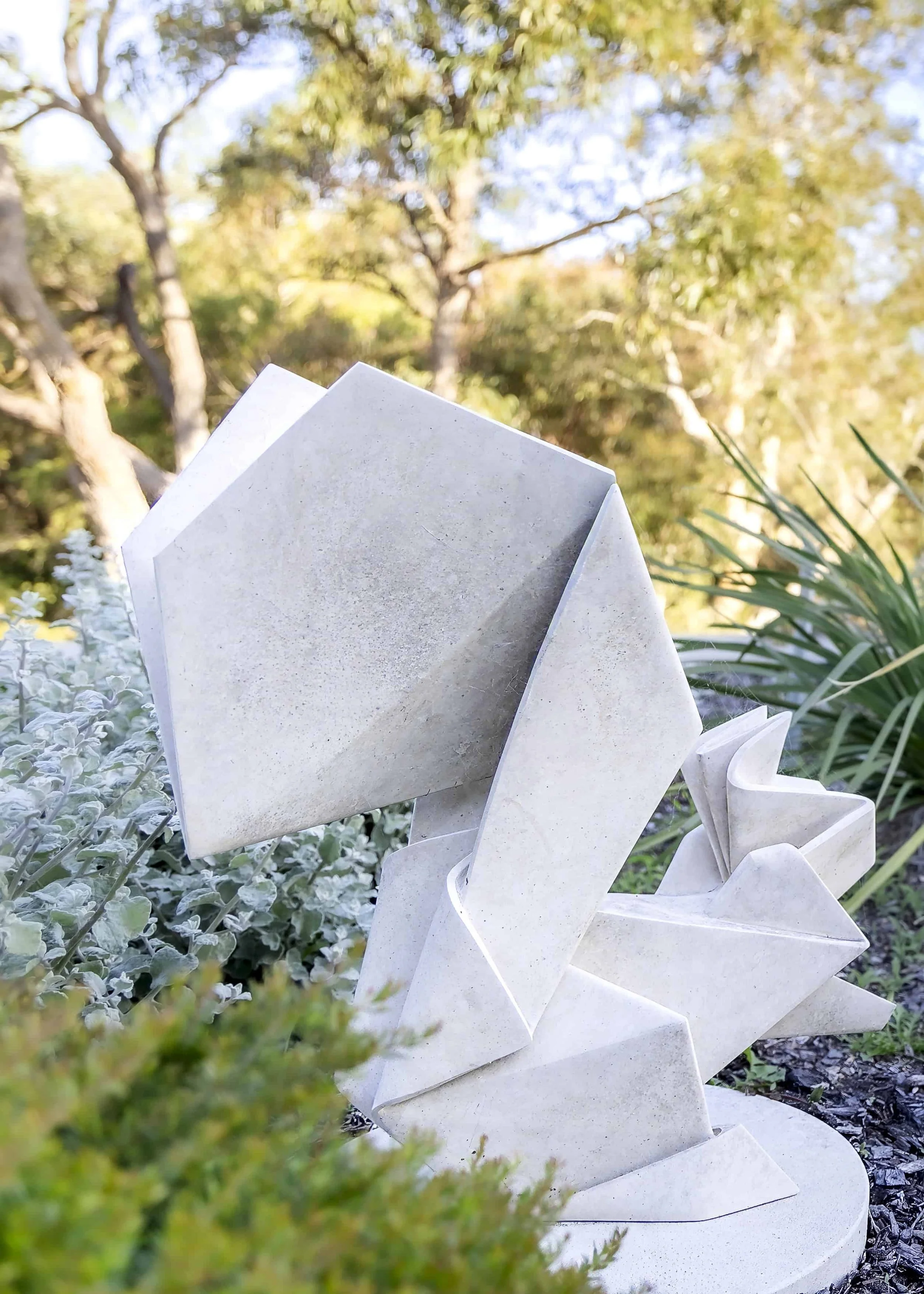 Abstract modern sculpture made of light-colored material, set outdoors among greenery and trees with a blurred background of tall trees and a partly cloudy sky.